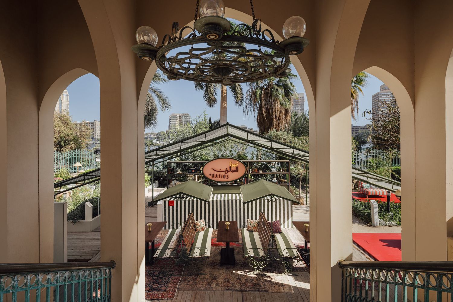 Elegant arched colonnade framing striped outdoor cafe pavilion with lush palm trees and city skyline beyond.