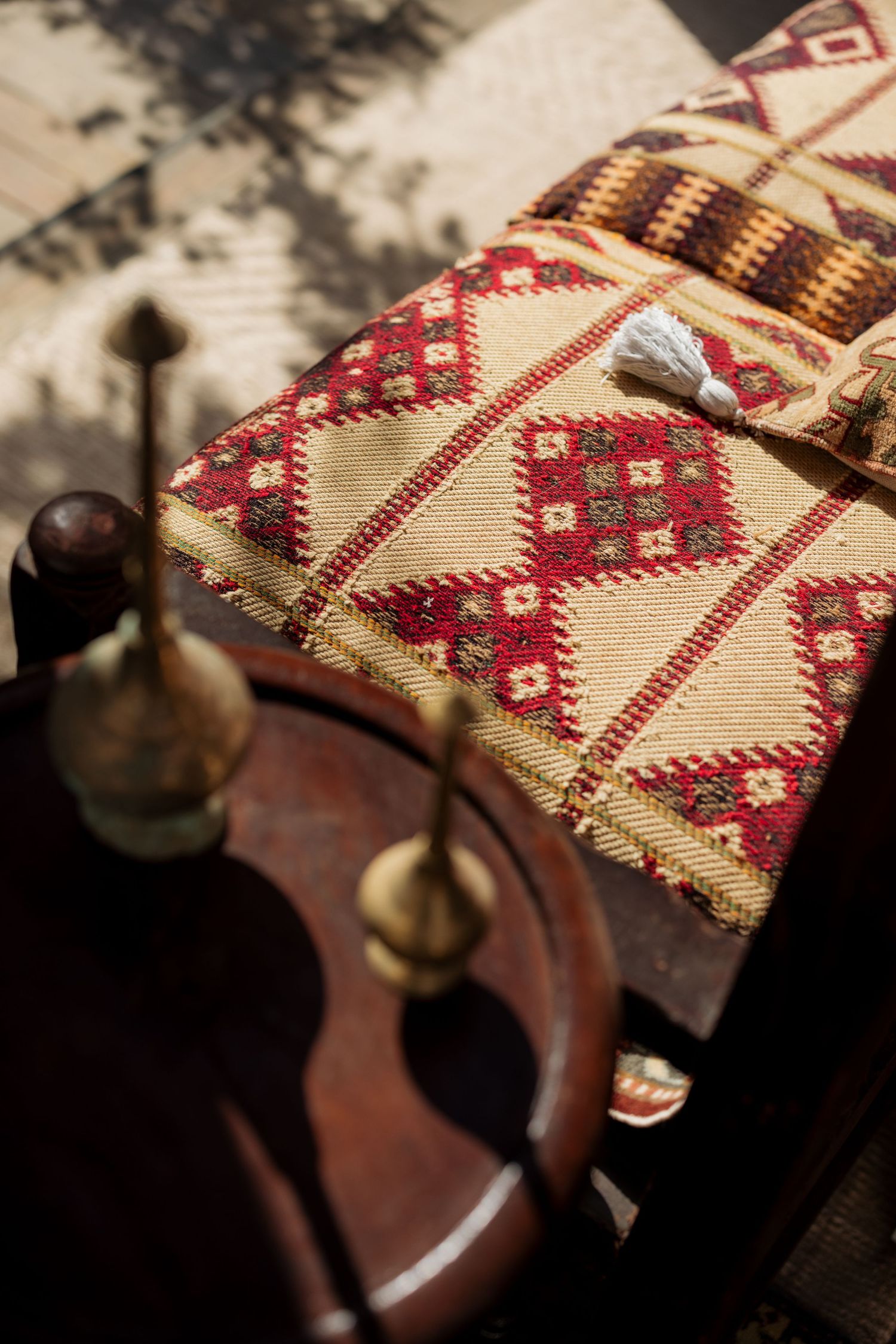 Traditional red and beige geometric patterned textile cushion with white tassel on wooden furniture in sunlight.