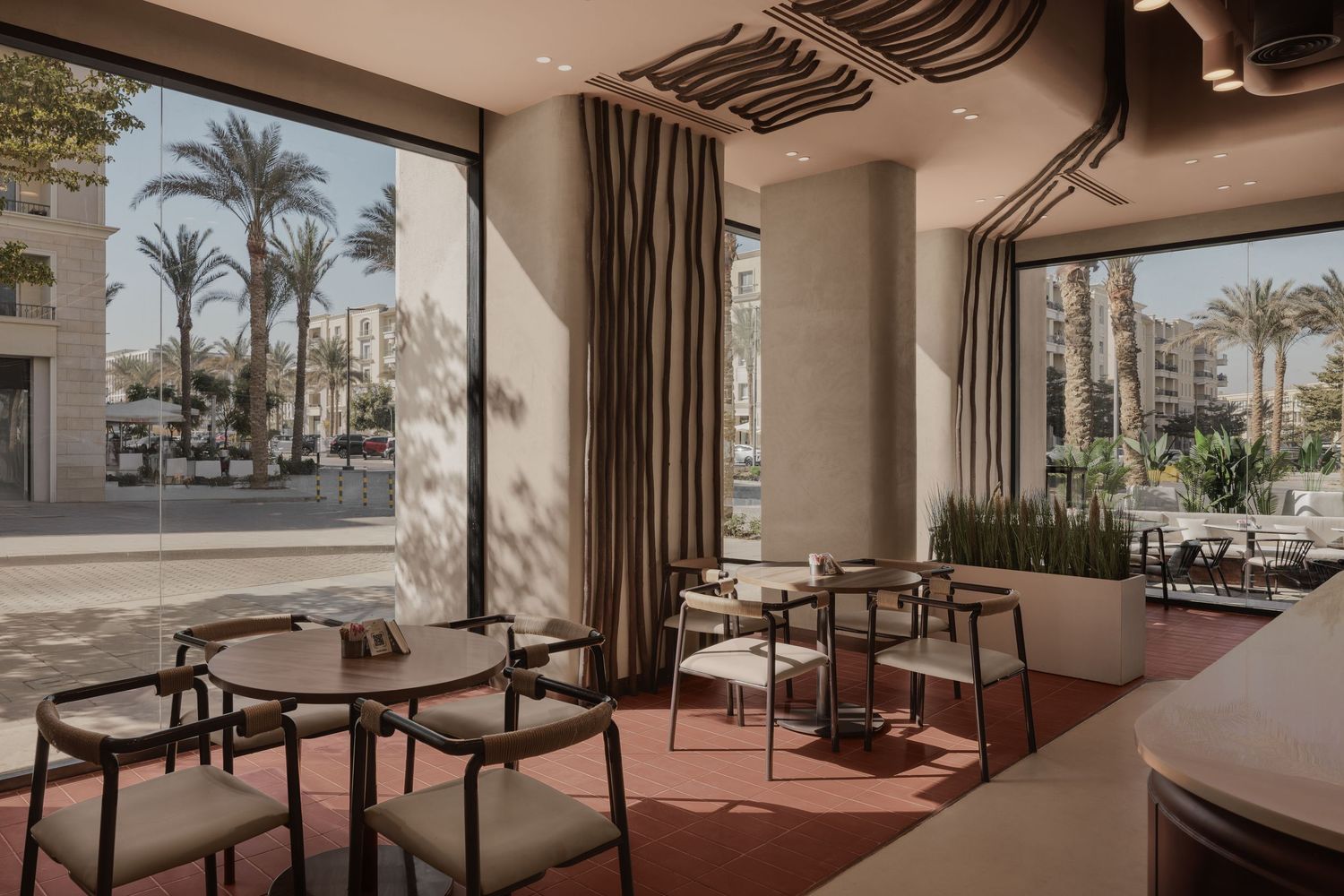 Stylish cafe interior featuring modern chairs, wooden tables, floor-to-ceiling windows, and palm tree views outside.