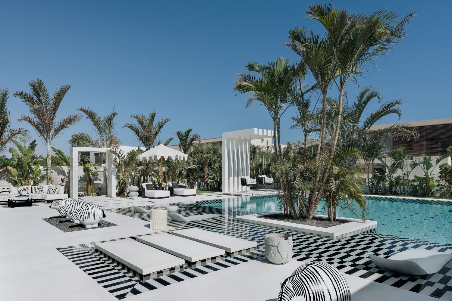 Luxury resort pool area with black and white checkered deck, white lounge chairs, and tropical palm trees under blue sky.
