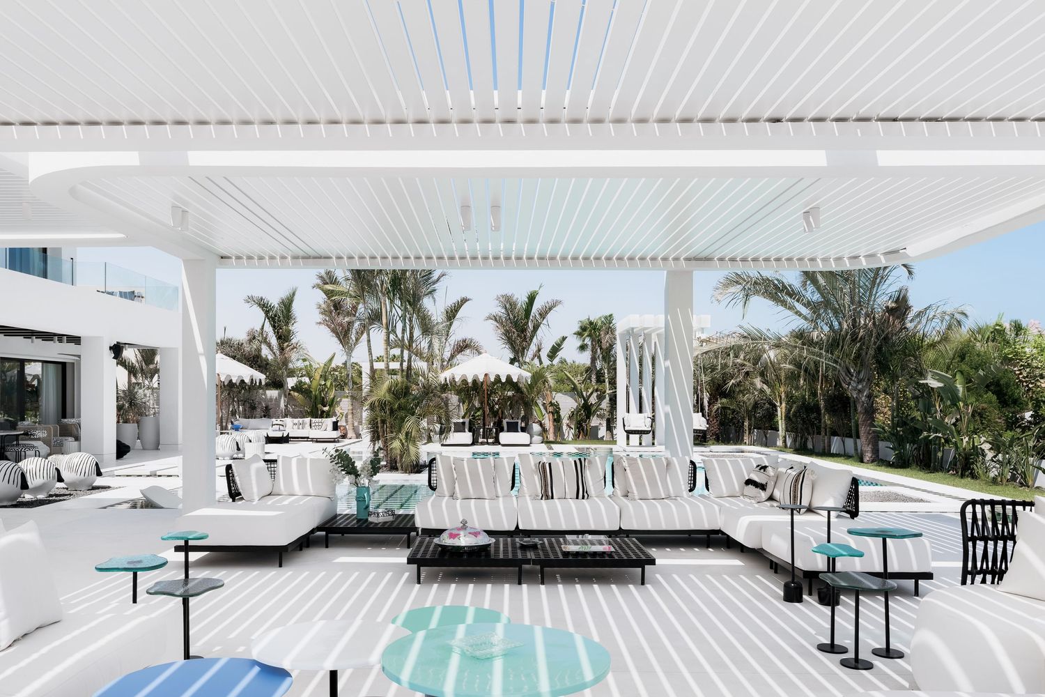 Luxurious white outdoor patio with striped sofas, pergola, turquoise accents, and tropical palm trees in a modern resort setting.