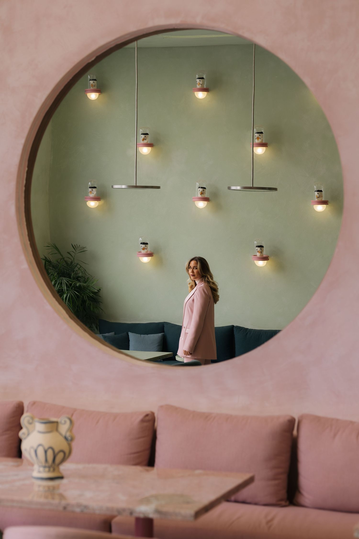 Elegant pink cafe interior featuring a circular mirror reflecting a woman and sage green wall with lights.