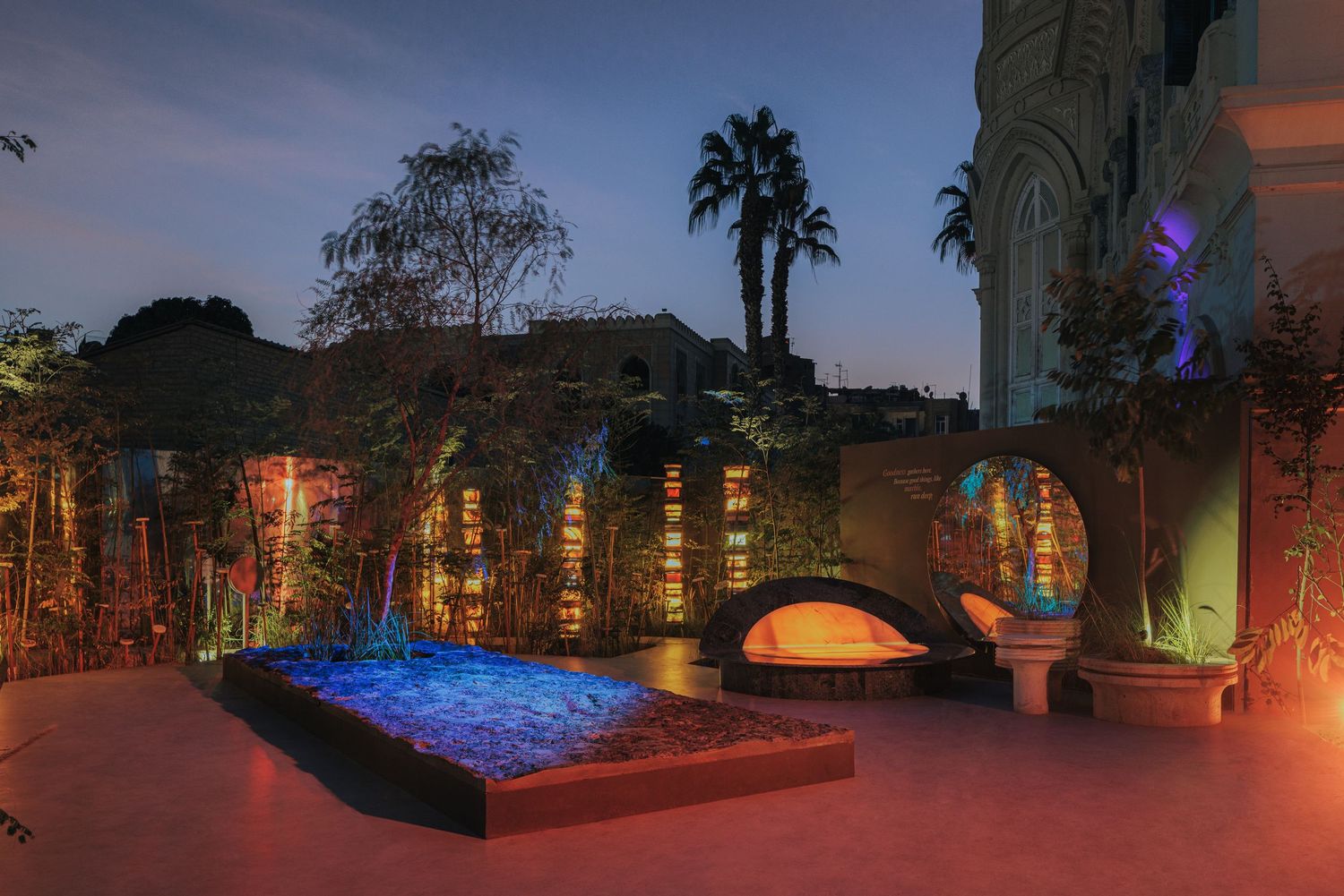 Night view of a colorful outdoor art installation with blue sand, arched benches, and illuminated bamboo.