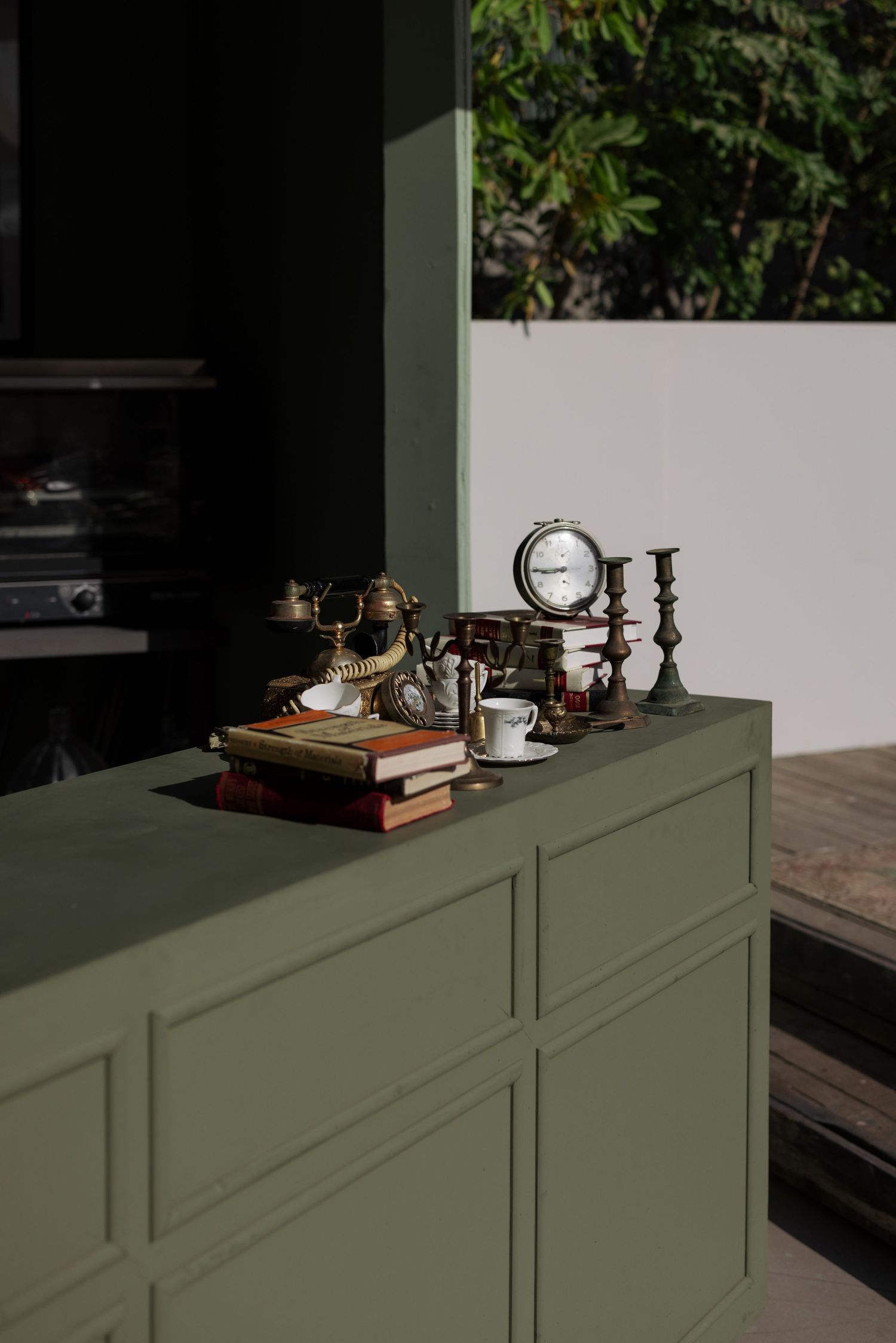 Olive green cabinet styled with vintage brass clock, candlesticks, telephone, and books in sunlit space.