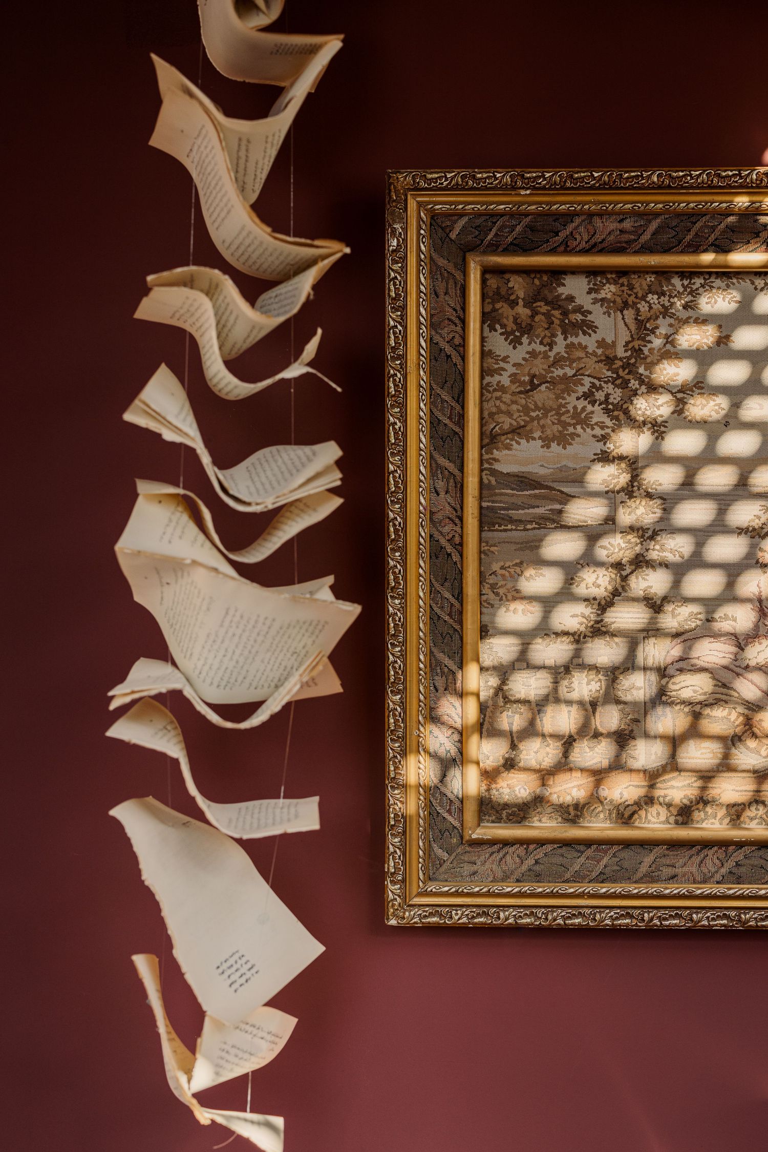Pages of an open book suspended on strings beside a gold ornate framed artwork on a dark red wall.