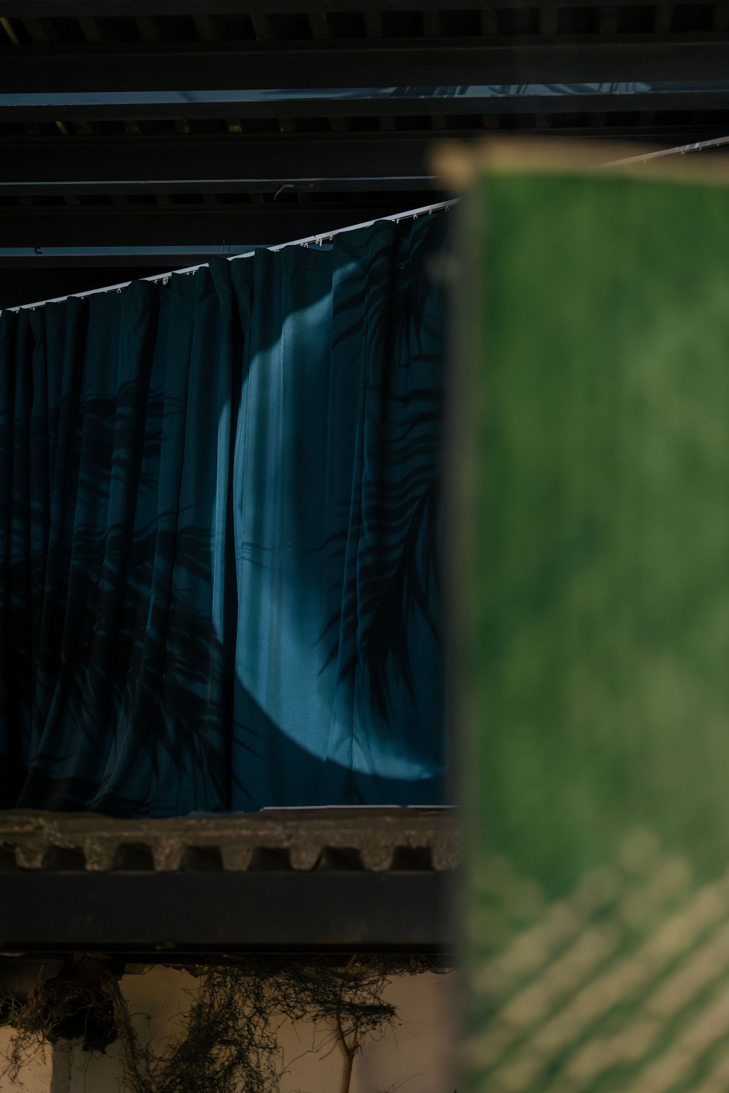 Teal curtain with tropical palm shadow print hanging beneath dark pergola beside green woven textile panel.