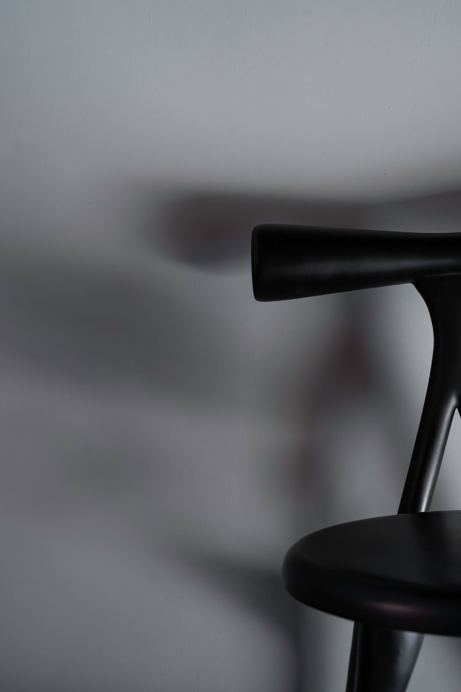 Black minimalist wooden chair back casting soft shadow on light gray wall in moody lighting.