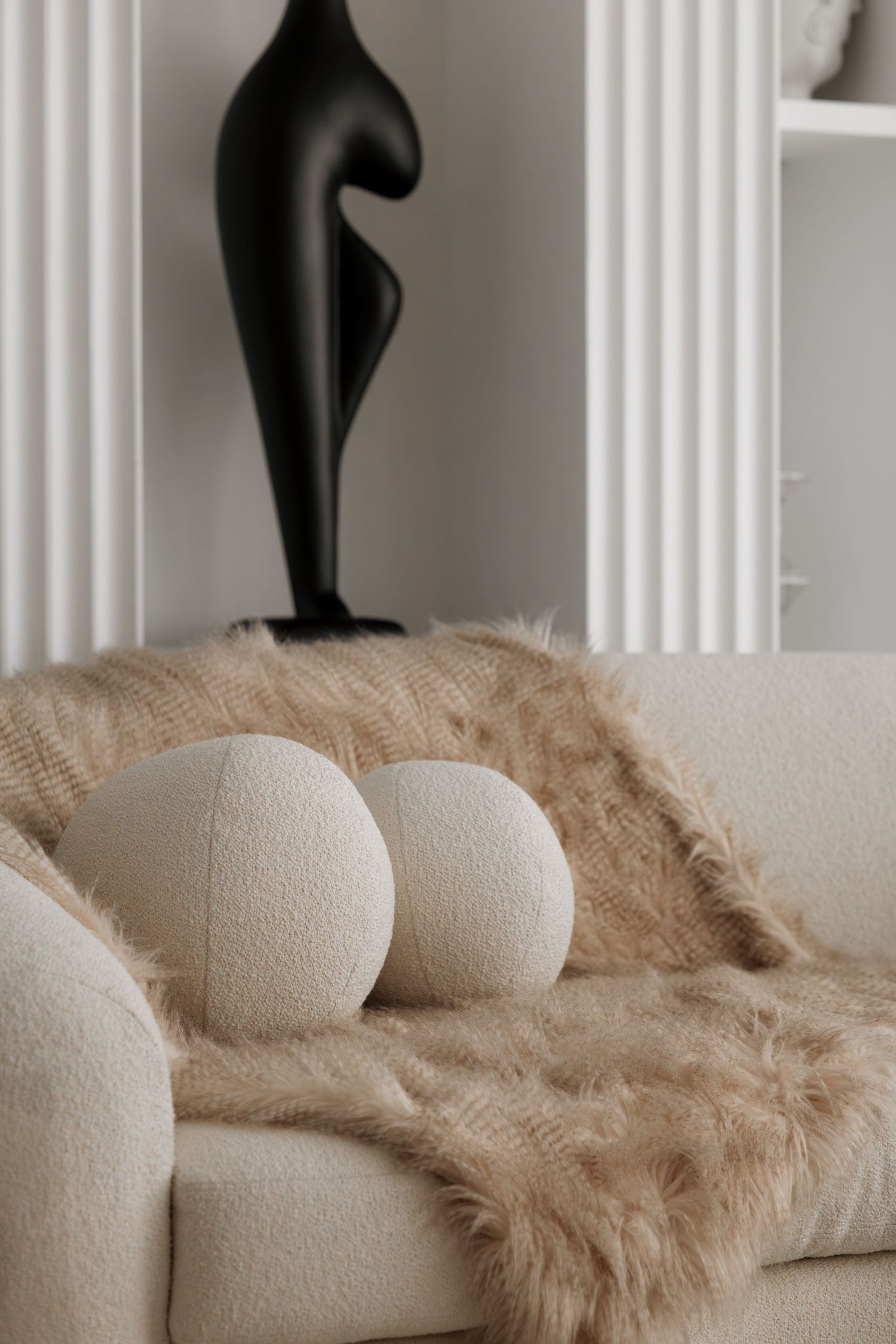 Close-up of white bouclé sofa with round pillows, faux fur throw, and black abstract sculpture in modern interior.