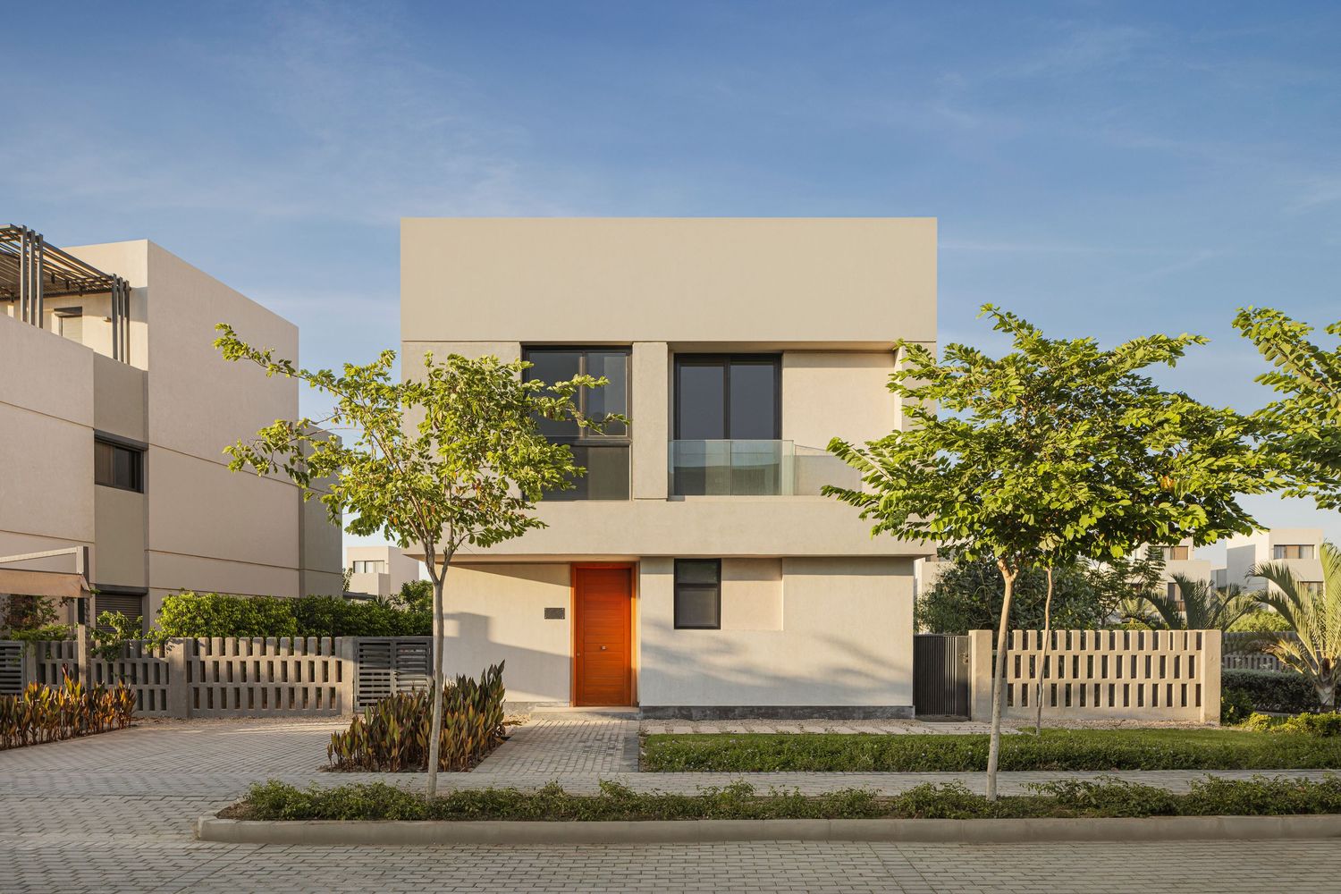 Modern two-story beige villa with orange door, large windows, lush trees, and paved driveway under a clear blue sky.