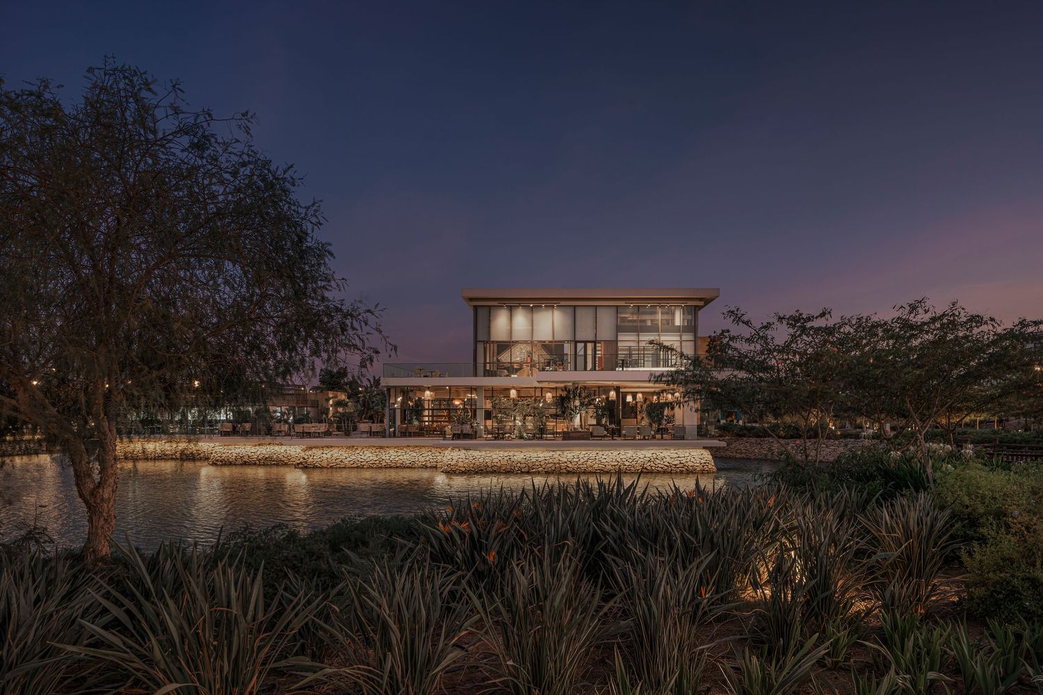 Elegant two-story building reflected in water at dusk with warm interior lighting.