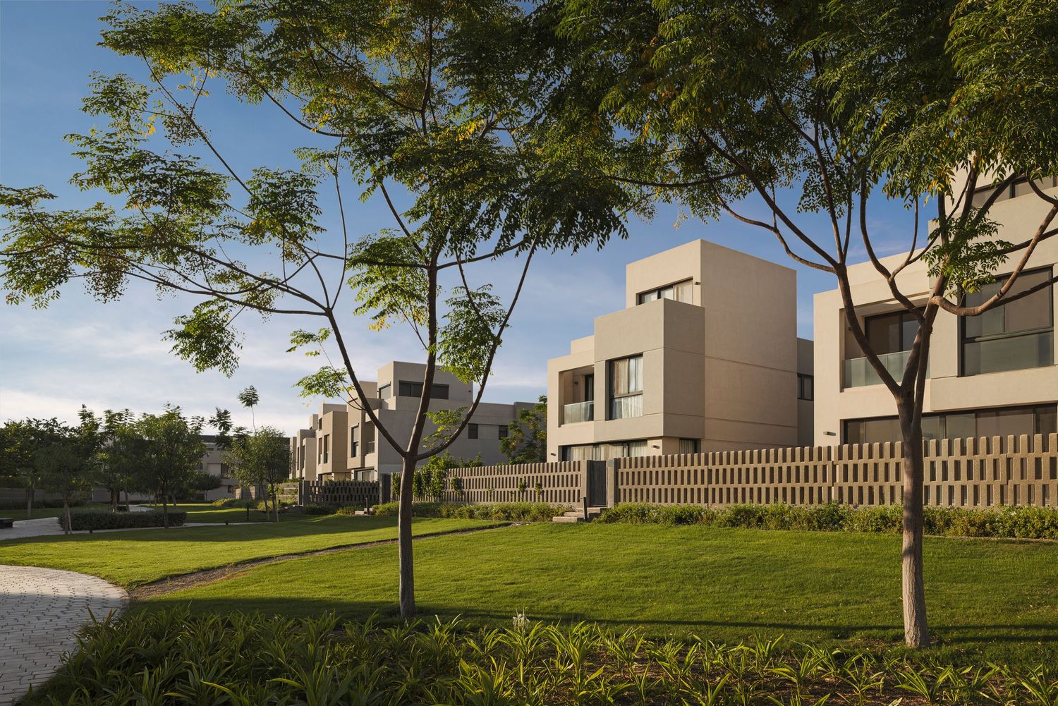 Modern luxury villas with beige facades surrounded by lush green lawns and trees under a clear blue sky.