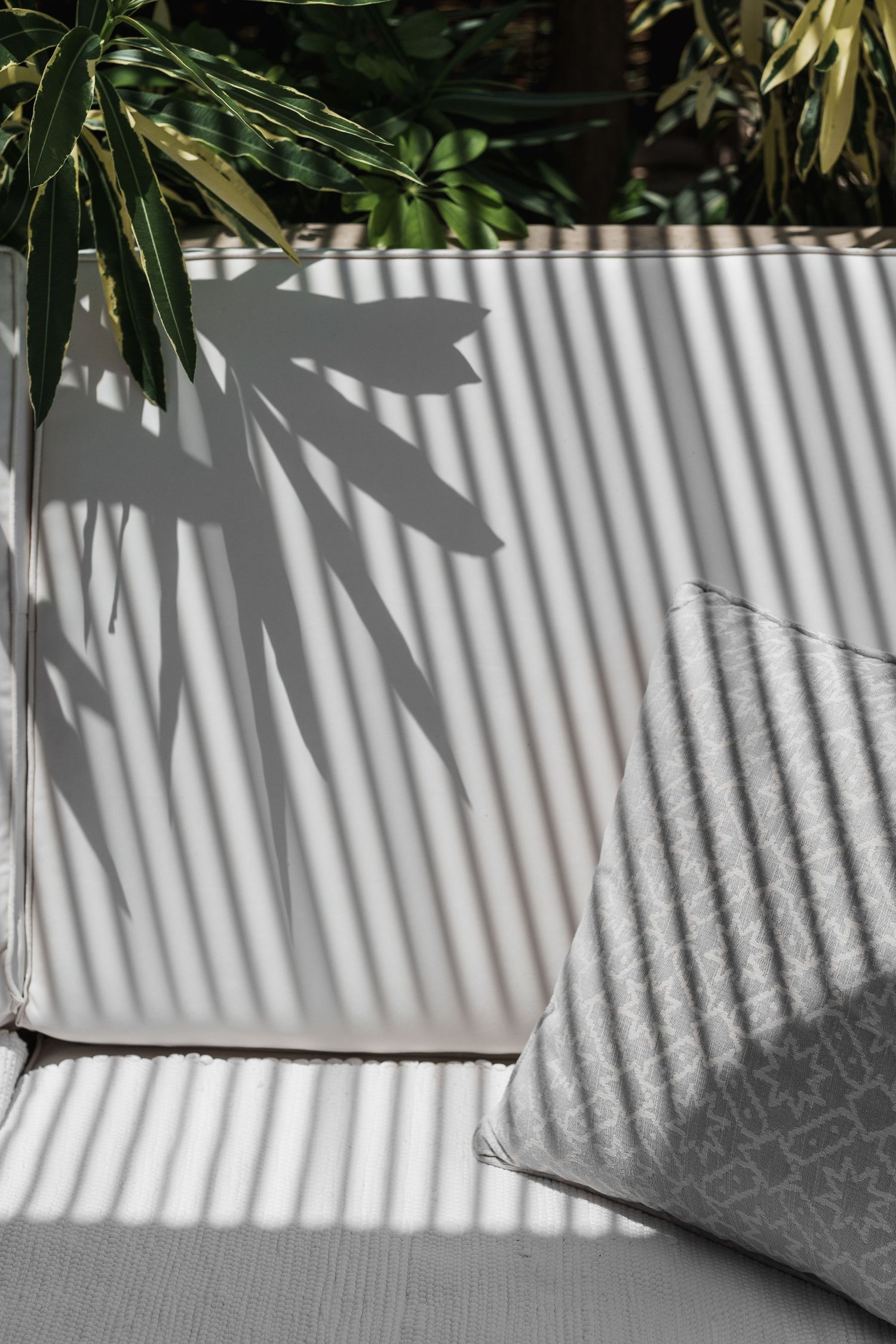 White outdoor sofa with geometric cushion casting striped shadows from tropical palm leaves in bright sunlight.