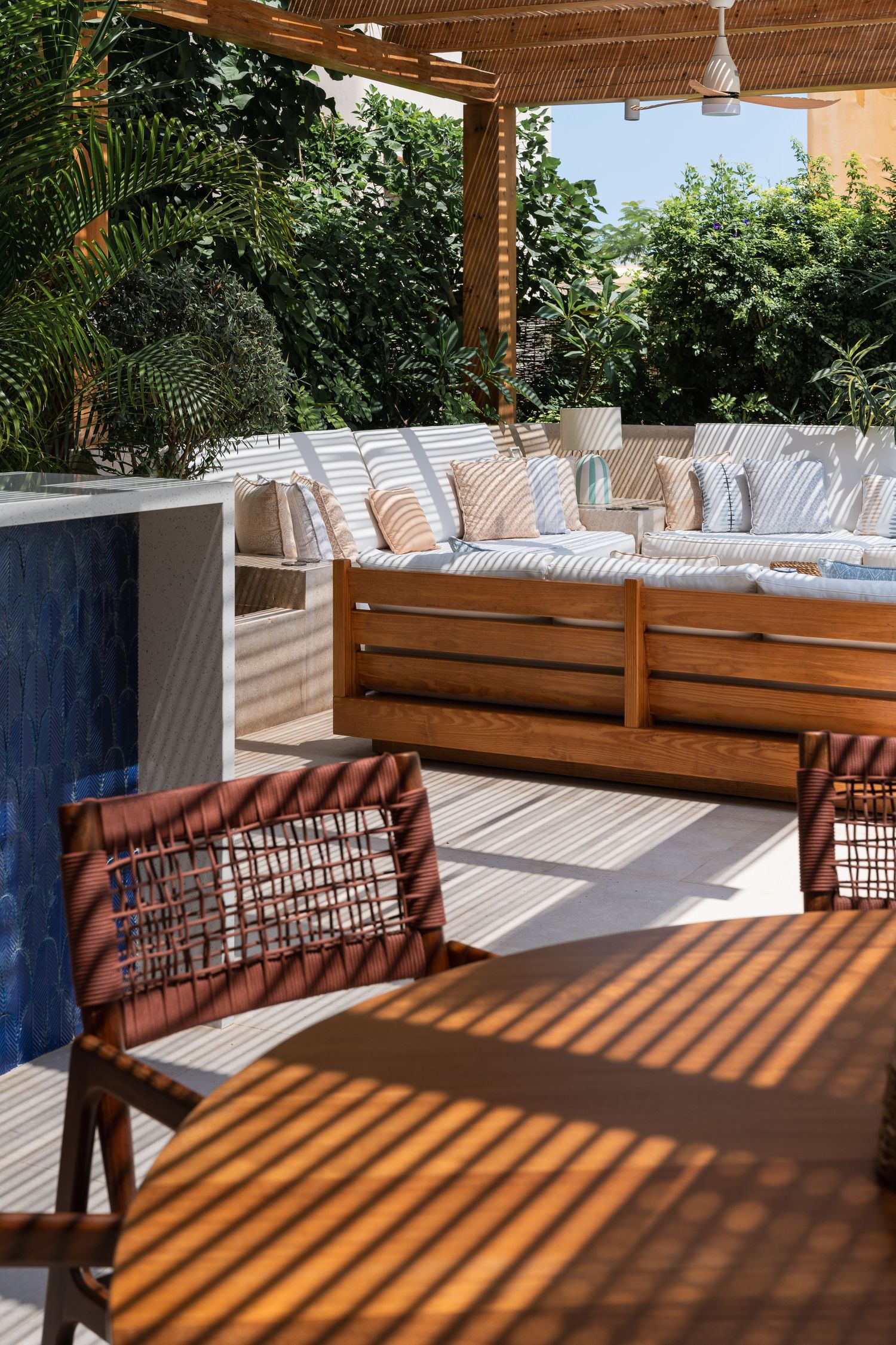 Sunlit outdoor terrace with wooden lounge furniture, striped cushions, pergola shade, and blue tiled pool edge.