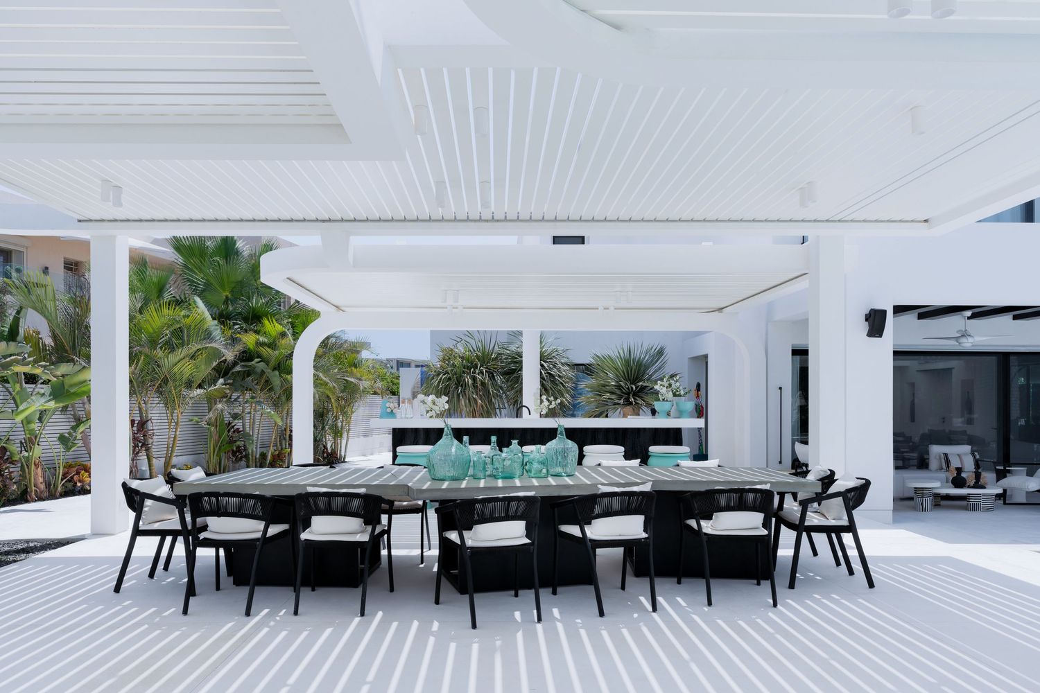 Outdoor dining area with black chairs around large table under white pergola with tropical pool setting.