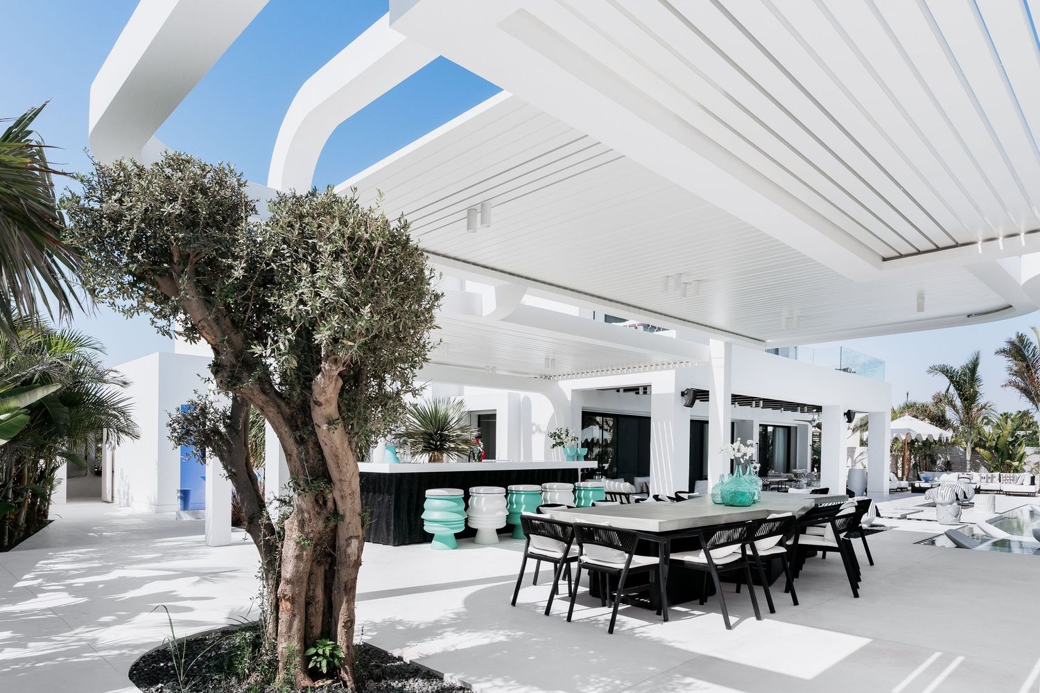 Luxurious white outdoor patio with black dining set, olive tree, turquoise accents, and pergola roof in modern villa.