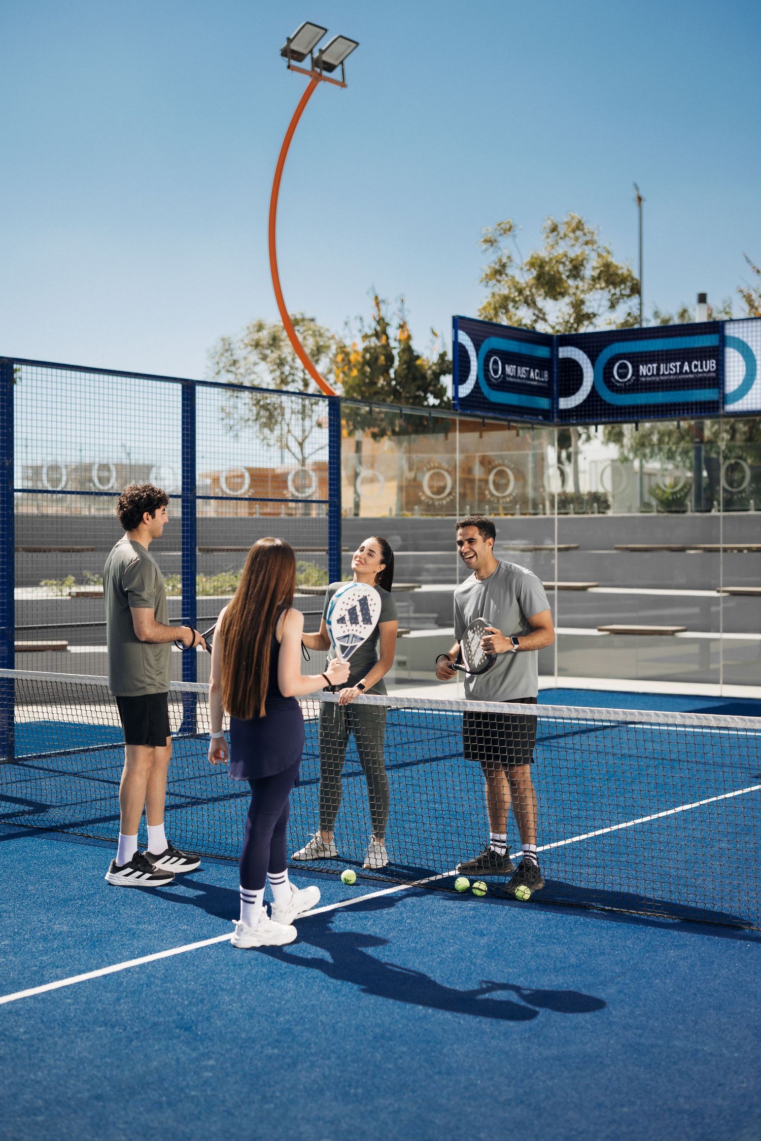 Four young adults socializing on a blue outdoor padel tennis court on a bright sunny day.
