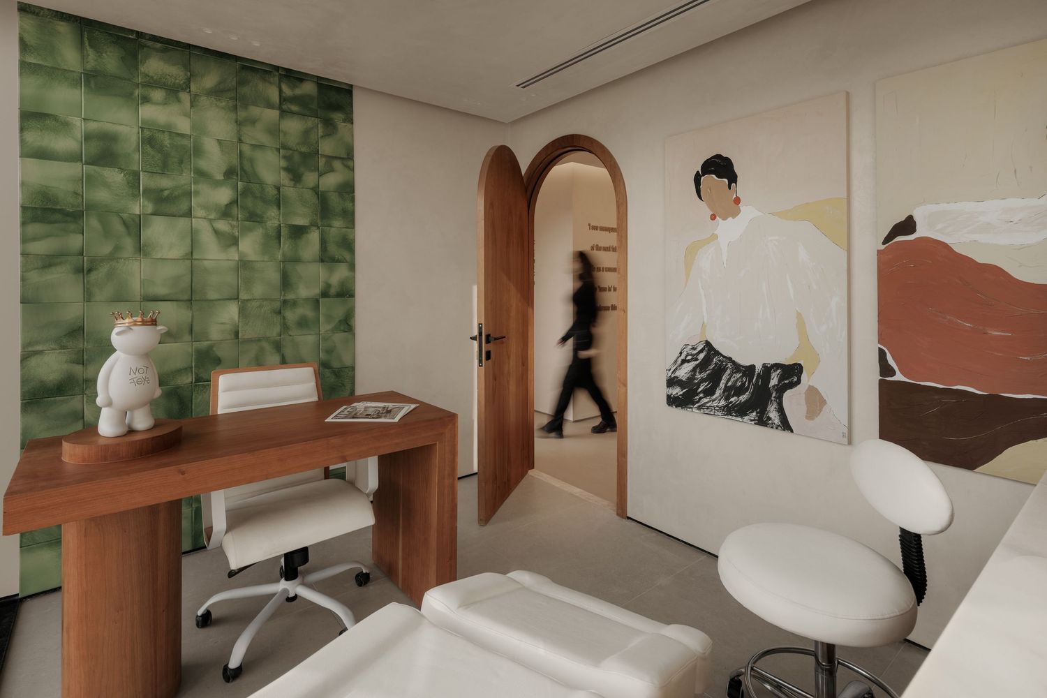 Modern treatment room with green tile accent wall, wooden desk, white chair, and large figurative wall art.