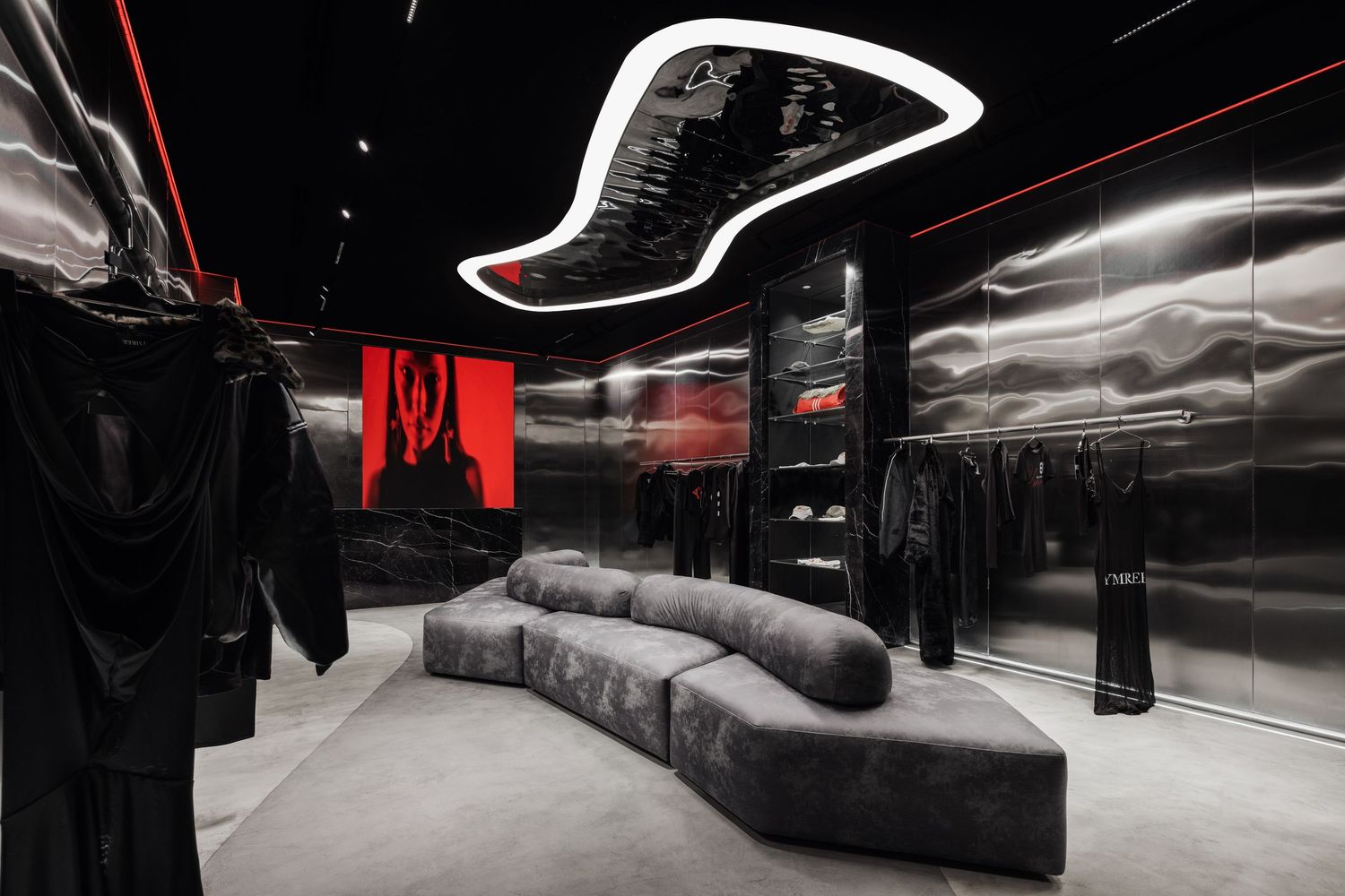 Dramatic fashion boutique with black metallic walls, red backlit display, curved gray sofa, and dark garments on hangers.