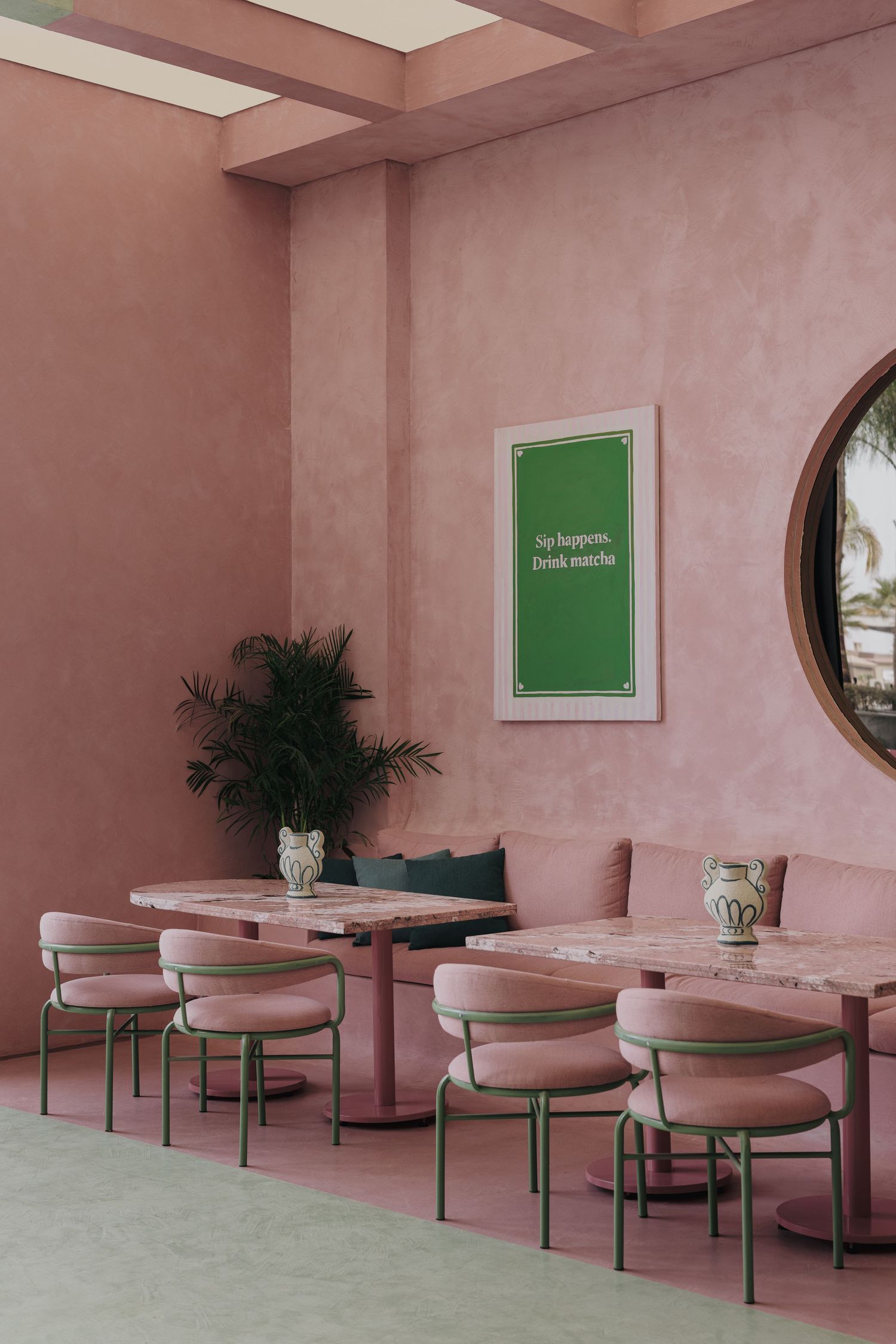 Pink cafe interior with green accents, marble tables, and a framed matcha quote poster on textured walls.