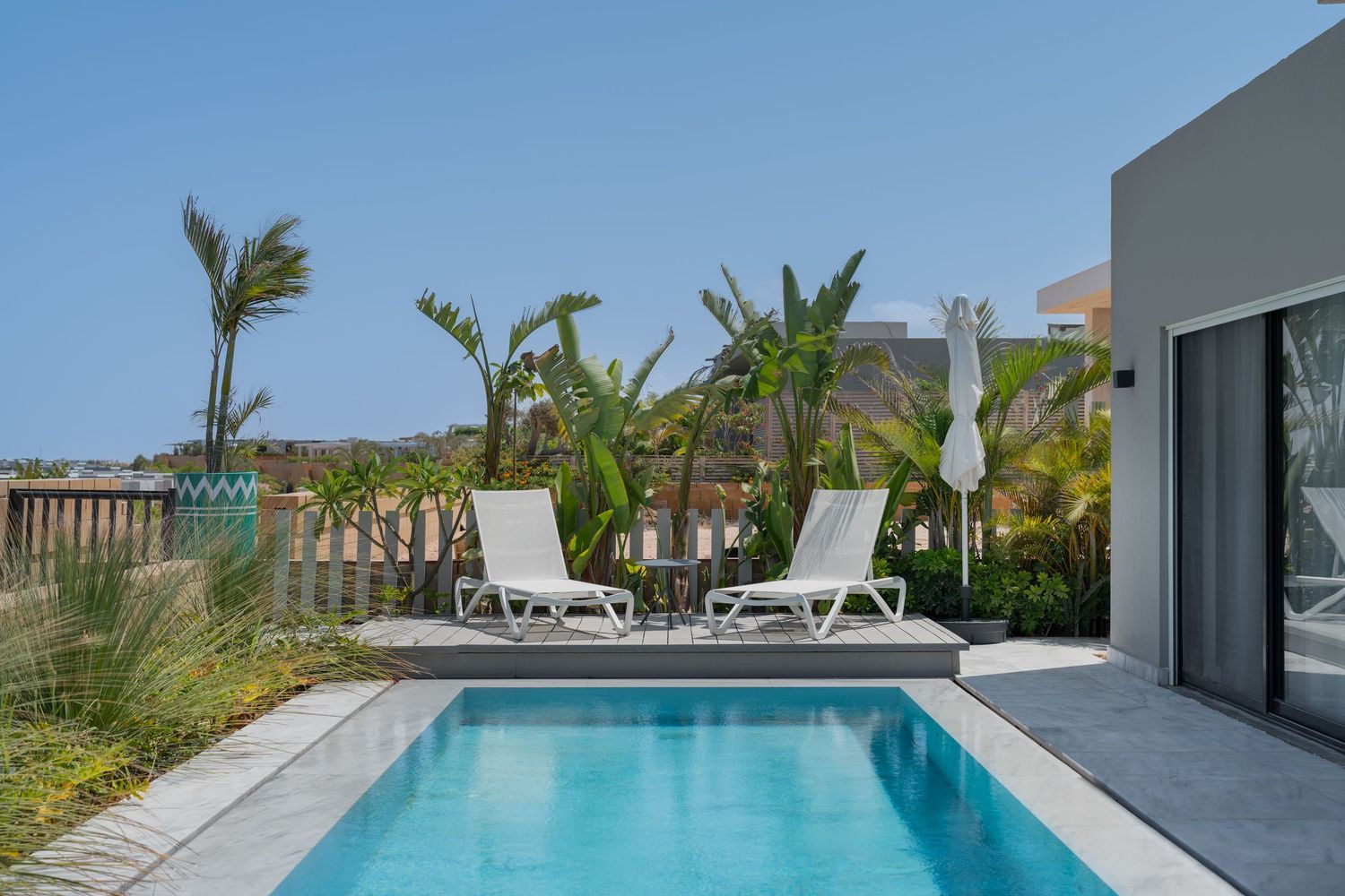 Luxury villa private pool with white lounge chairs, tropical landscaping, and clear blue sky backdrop.