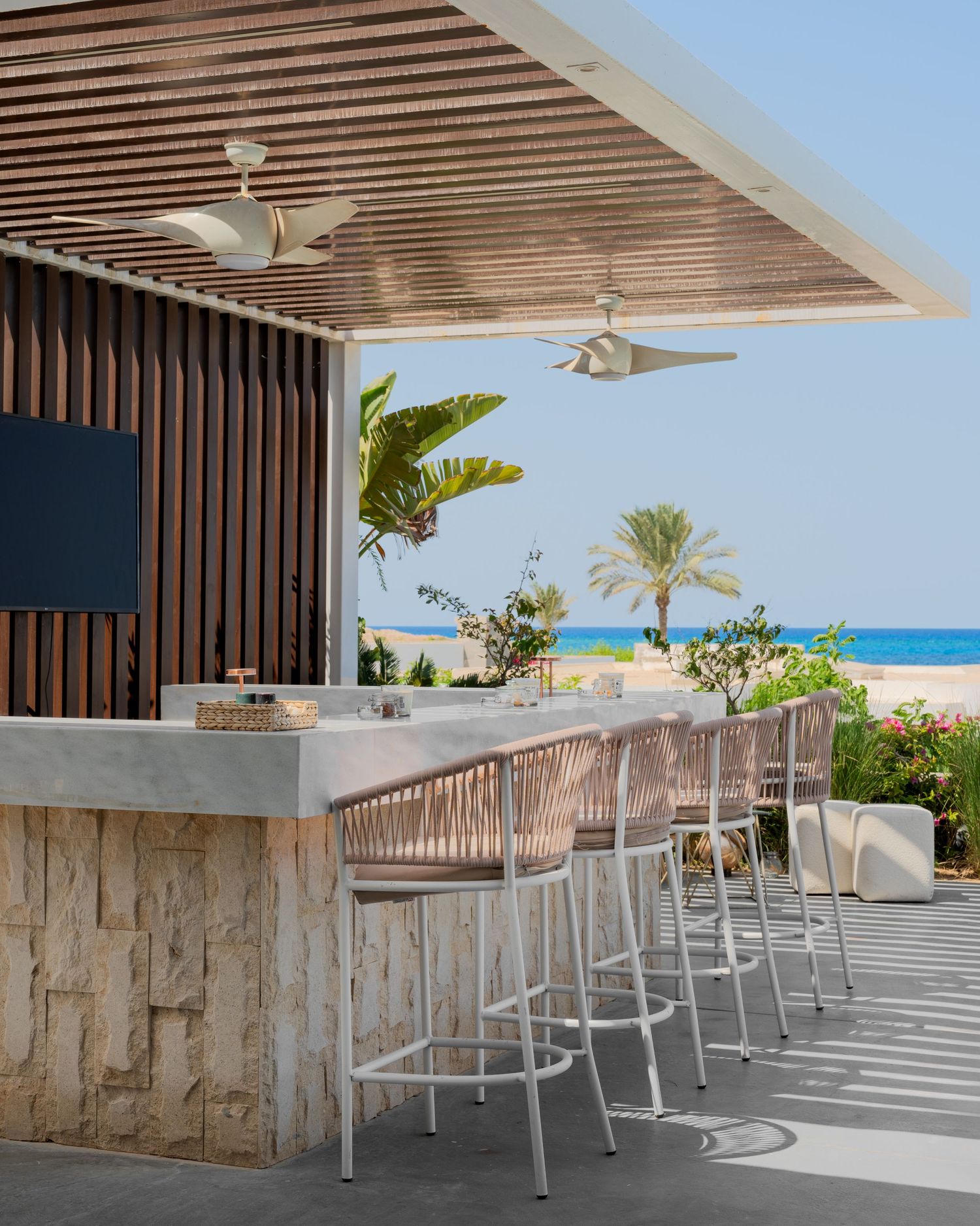 Beachfront outdoor bar with stone counter, woven bar stools, slatted wood pergola, ceiling fans, and palm tree ocean views.