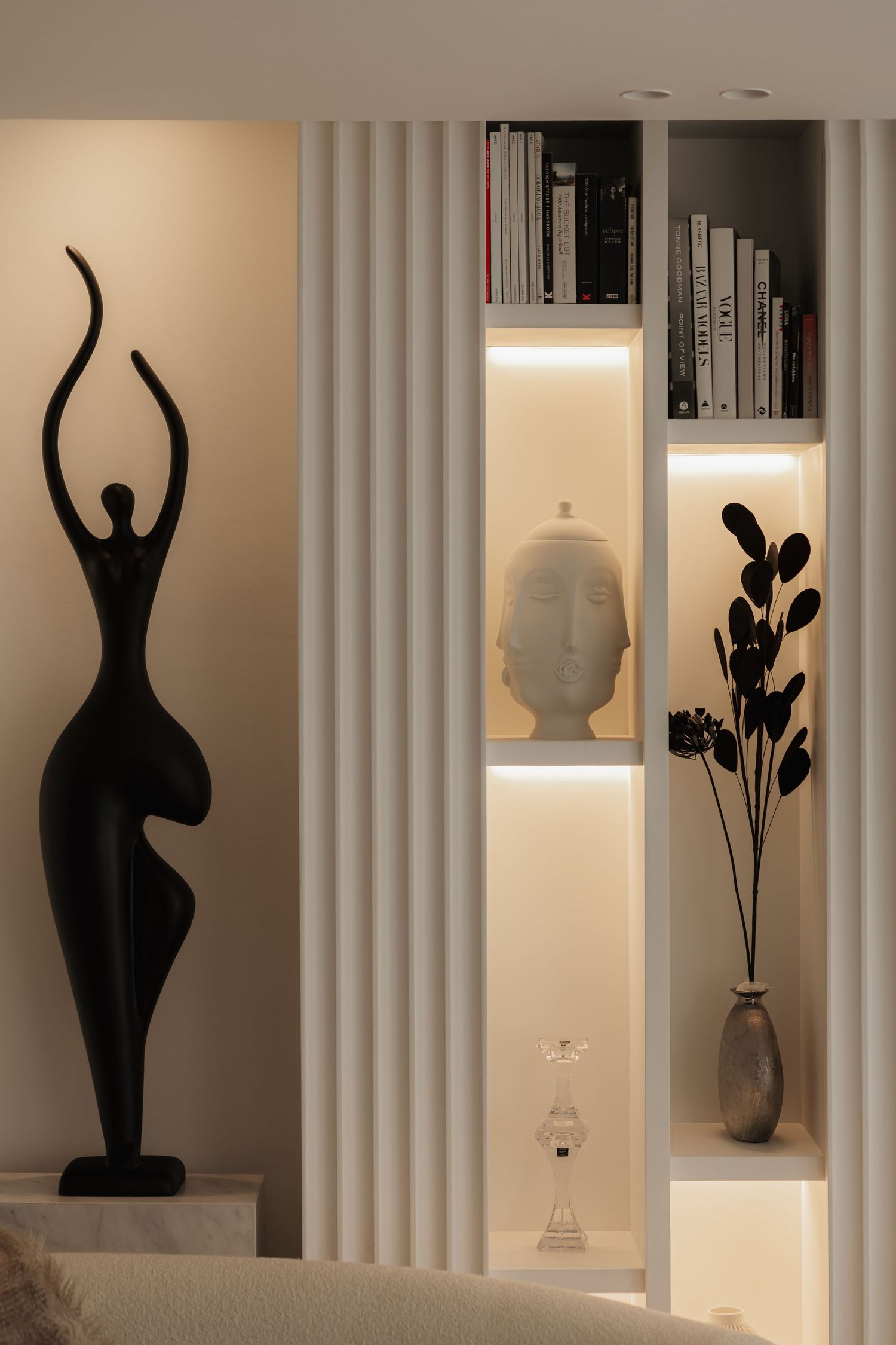 Stylish interior vignette with tall black female figurine sculpture, illuminated built-in shelves, and decorative objects.