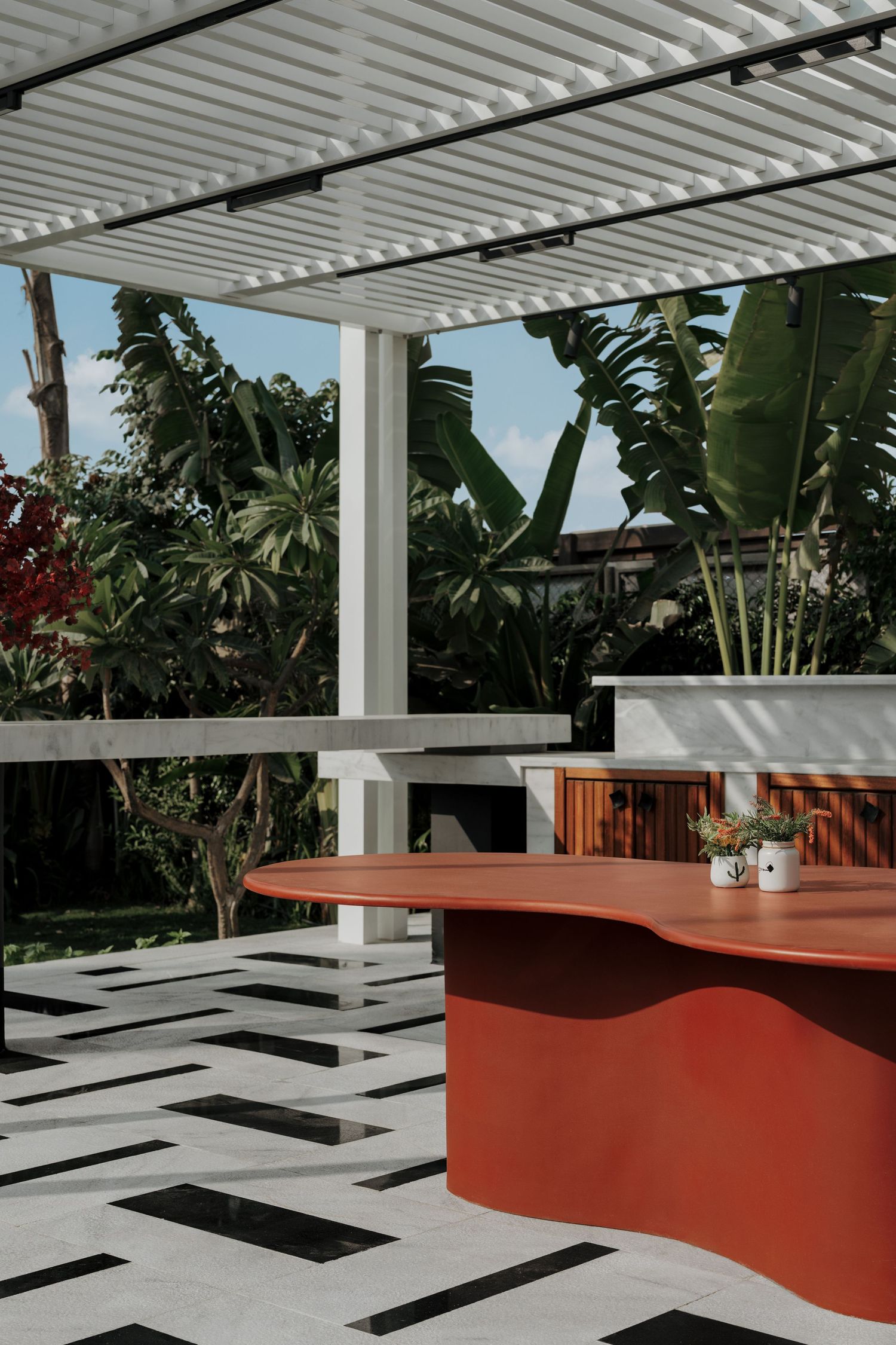 Stylish outdoor pergola with red curved bar counter, tropical garden backdrop, and geometric tile floor.