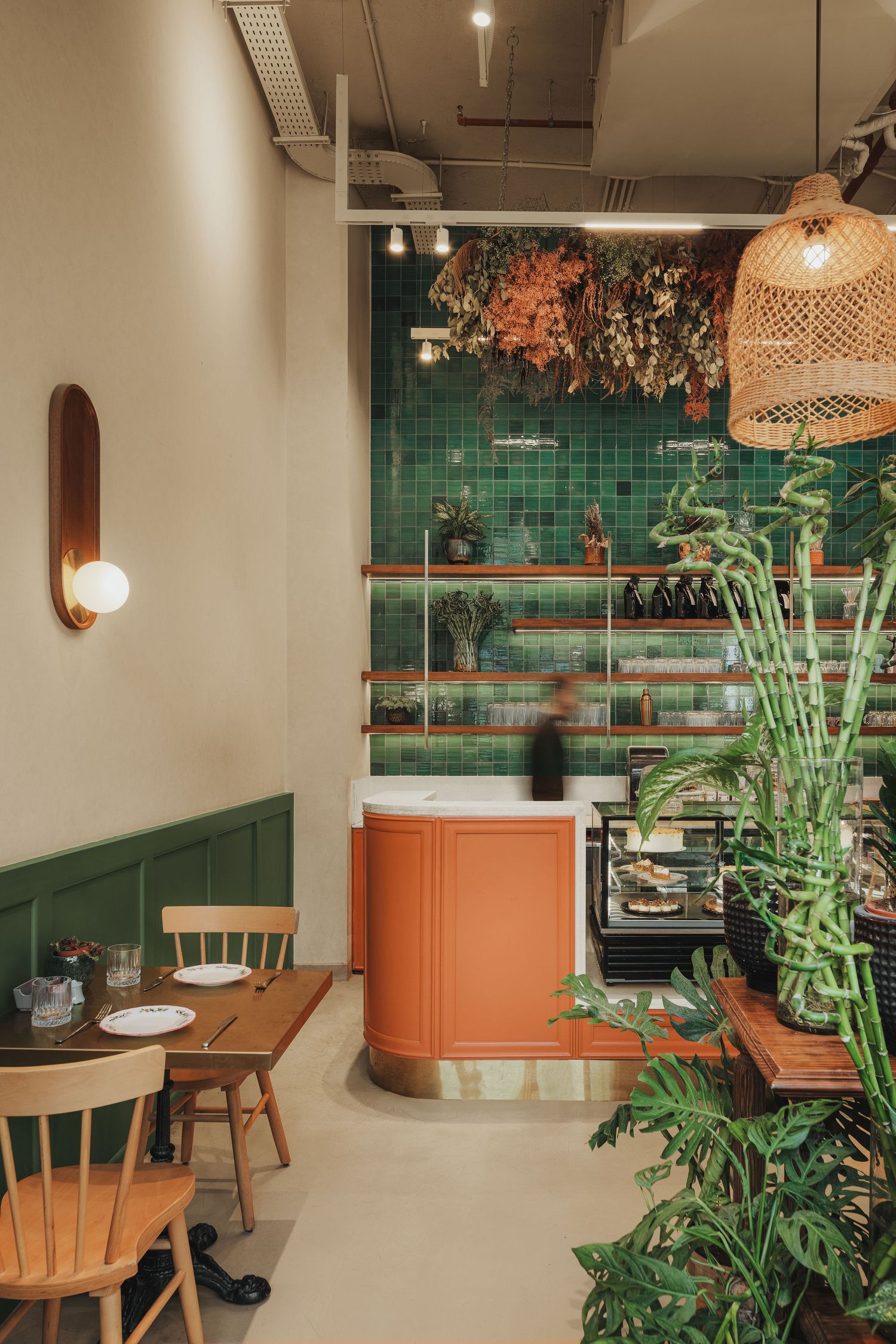 Cozy cafe interior with green tile wall, terracotta counter, wooden shelves, lush plants, and rattan pendant light.