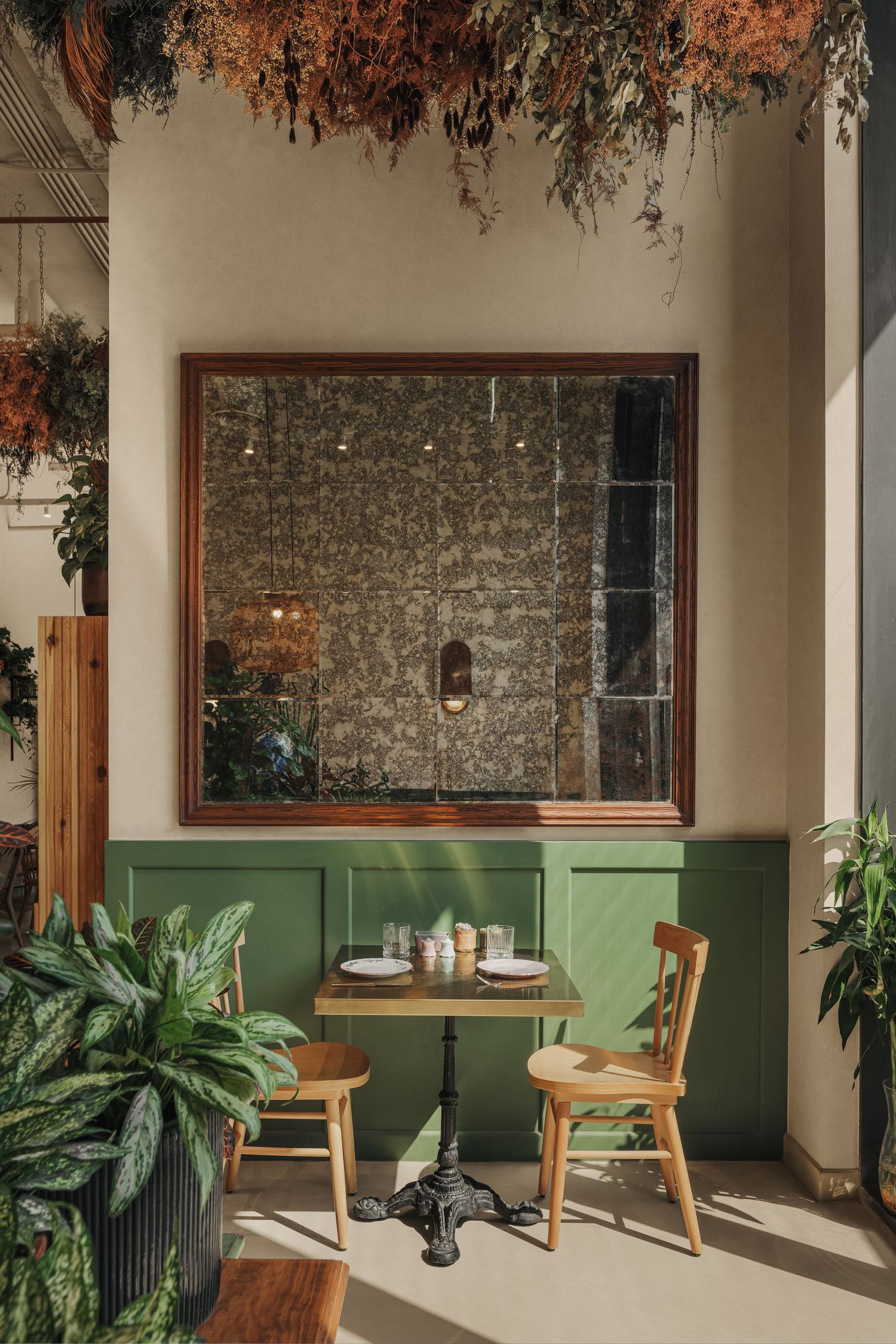 Cozy cafe dining nook with green wainscoting, wooden chairs, bistro table, large mirror, and lush hanging dried botanicals.