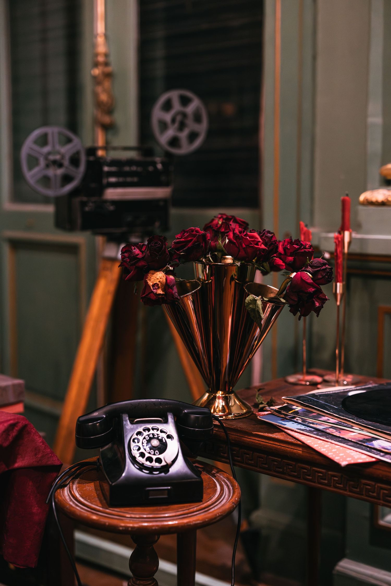 Antique black rotary telephone beside gold vase with dark red roses and vintage film reel on table.