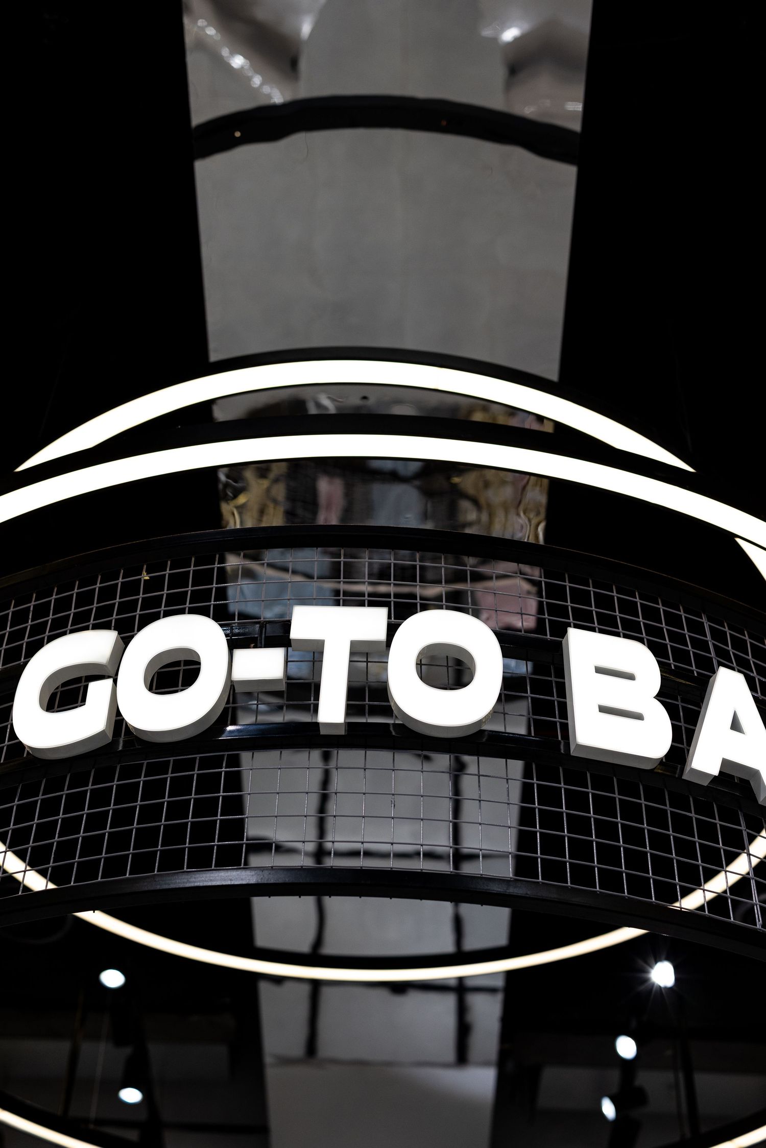 Illuminated circular GO-TO BA retail store sign with white LED lettering on dark black mesh background.