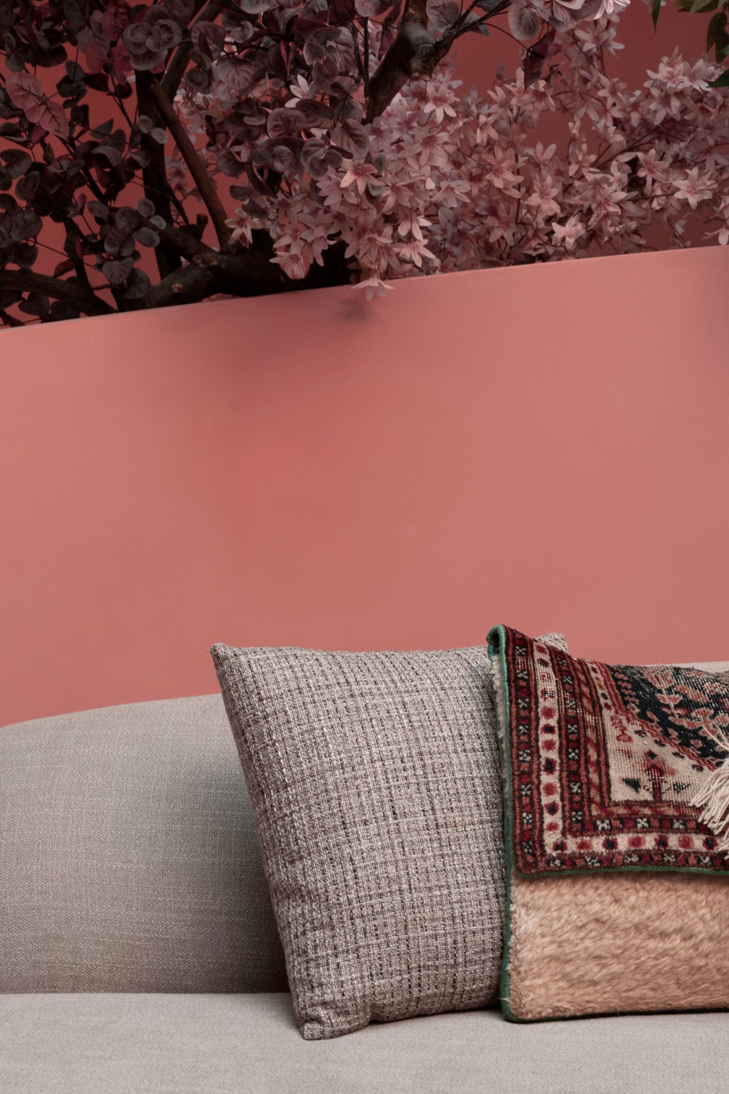 Grey textured sofa with woven and patterned cushions against a dusty pink wall with dark floral branches above.
