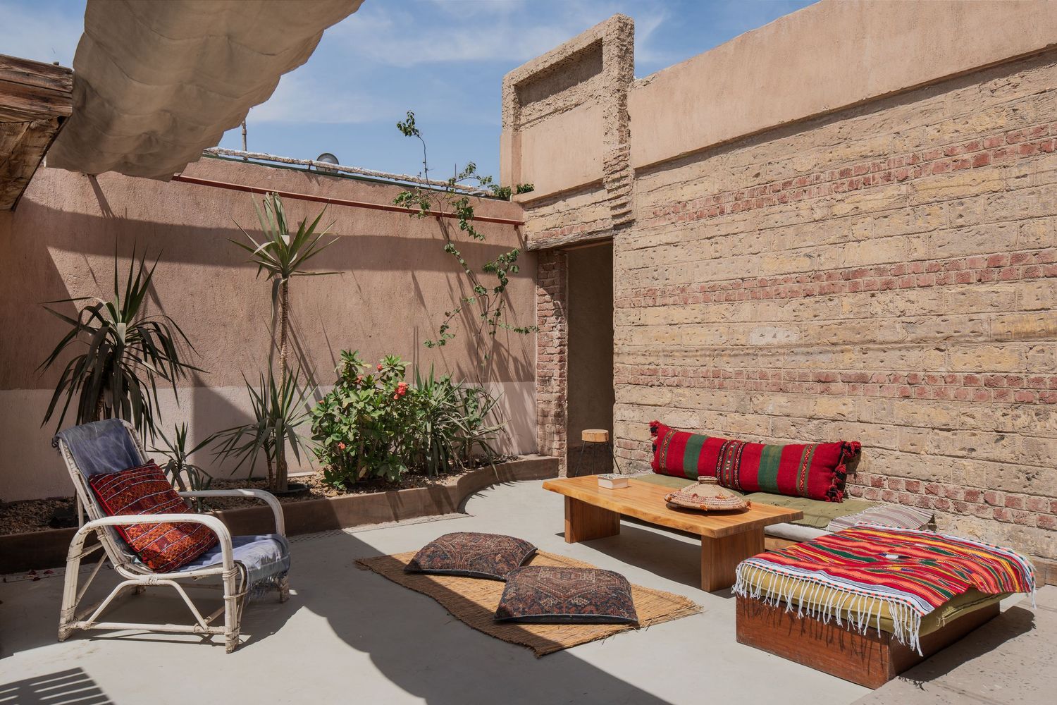 Sun-drenched Moroccan courtyard patio with rattan chair, kilim cushions, low wooden table, and terracotta walls.