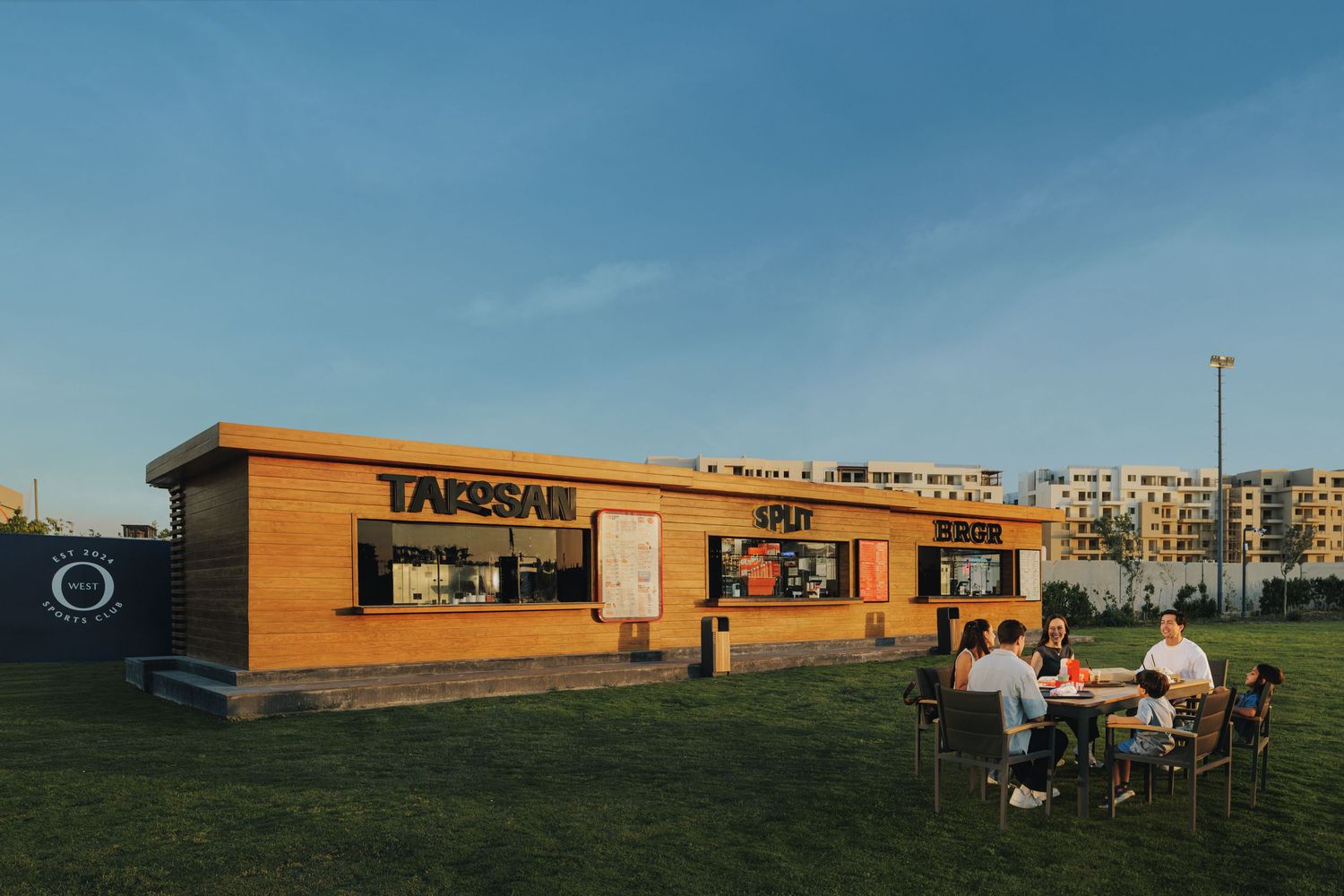 Outdoor food kiosk with takeaway and split signs where guests dine at tables on a green lawn at sunset.