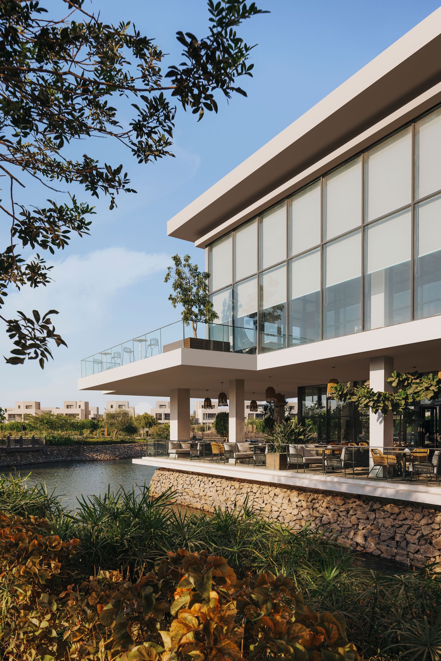 Modern waterfront building with large glass windows, outdoor terrace seating, and lush tropical greenery under a clear blue sky.