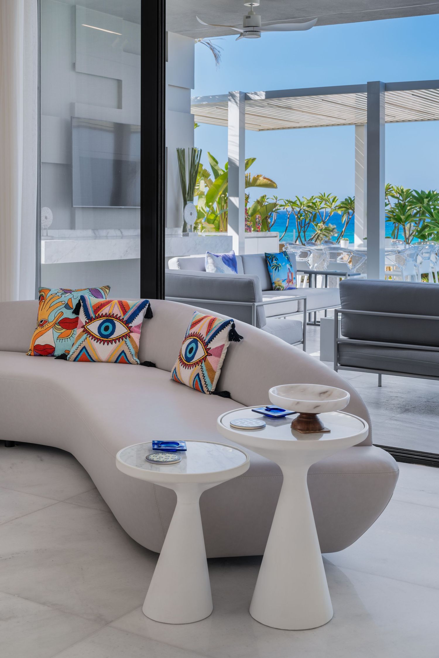 Curved white sofa with colorful eye-print pillows, marble side tables, and open view of tropical poolside terrace.