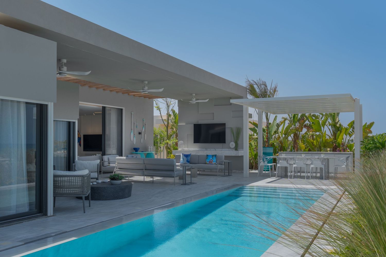 Modern villa outdoor living area featuring a rectangular pool, covered lounge space, and lush tropical garden.