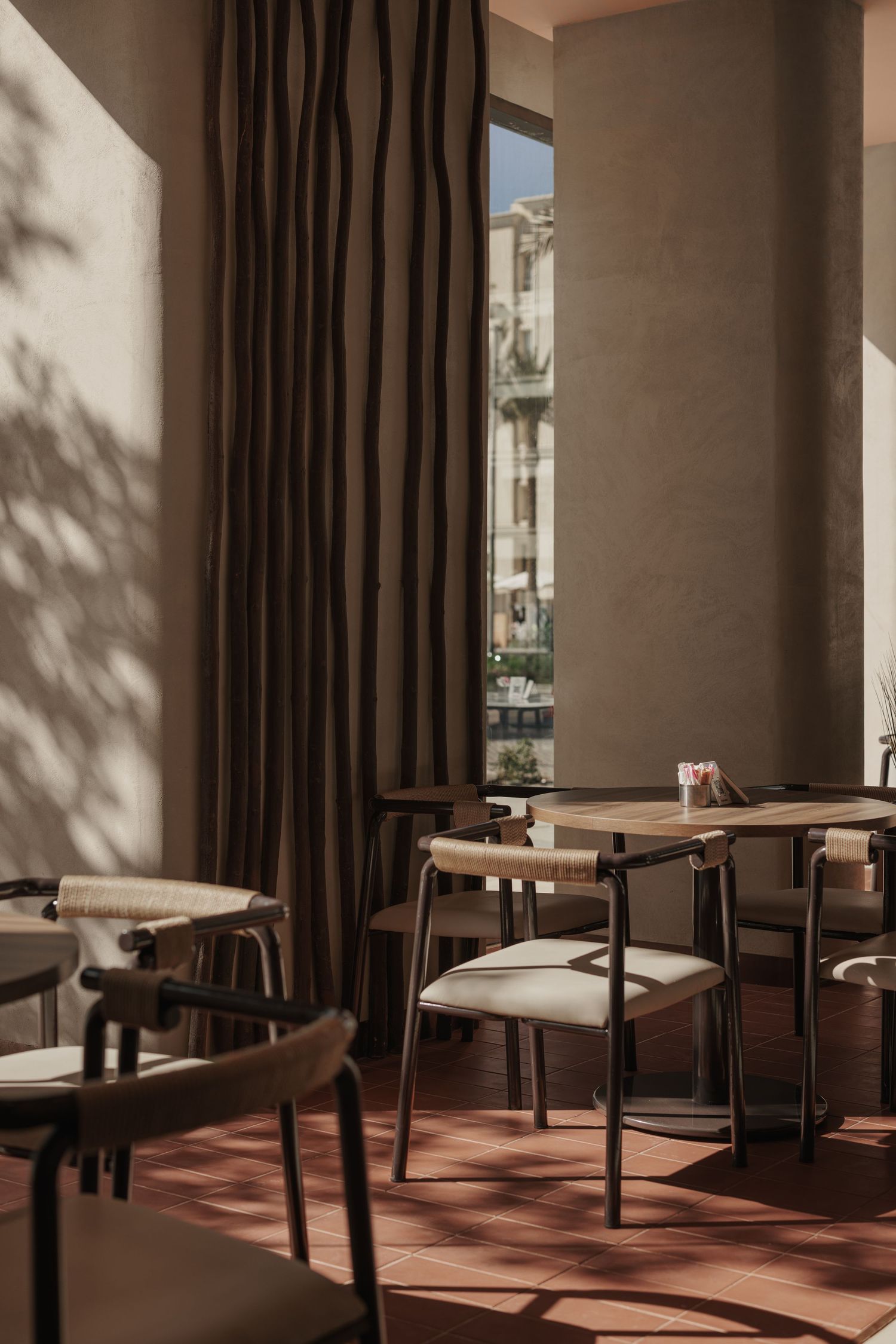 Cozy cafe seating area with woven chairs, wooden tables, terracotta floor tiles, and large window with curtains.