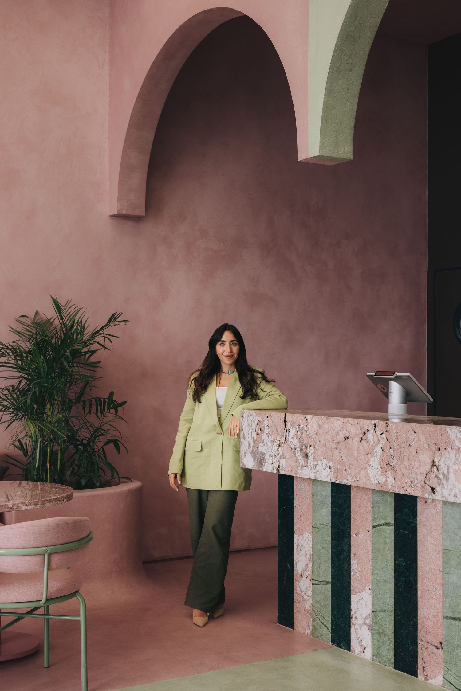 Stylish woman in light green blazer posing near pink travertine reception desk inside pink arched architectural interior.