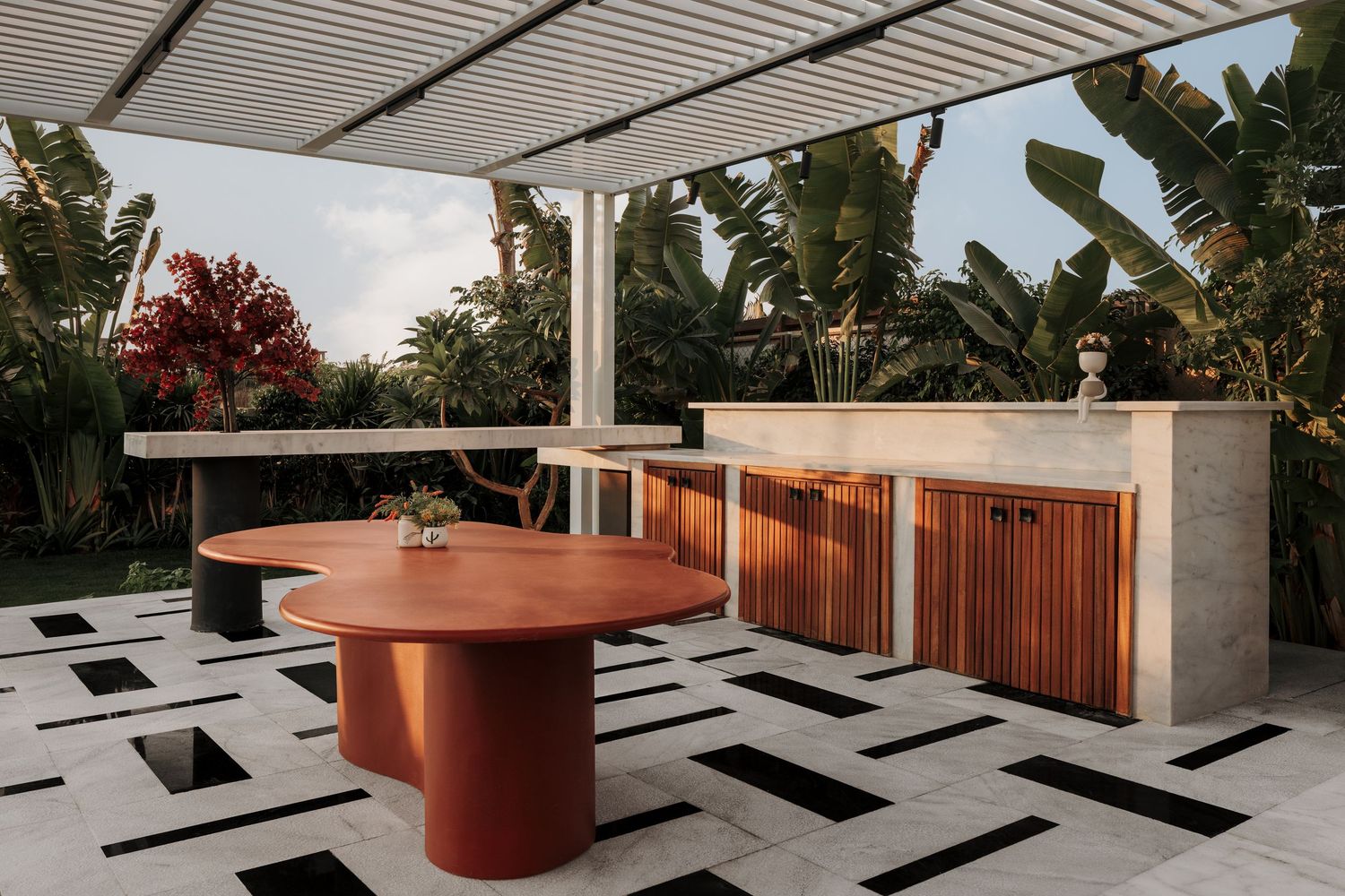 Contemporary outdoor dining area with red organic-shaped table, geometric tile floor, and wood cabinetry.