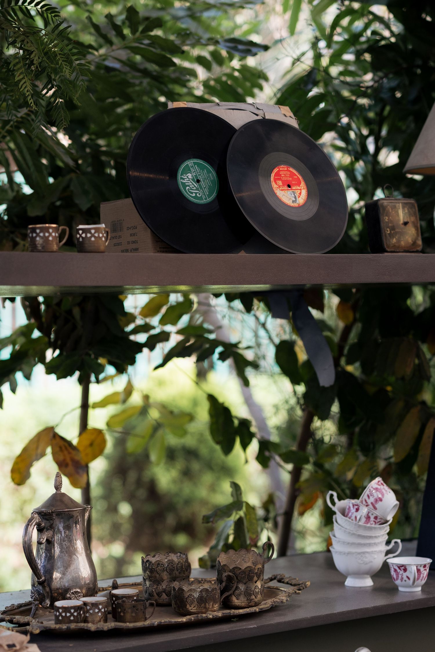 Two vintage vinyl records displayed on shelf with silver tea set and floral cups against green foliage.
