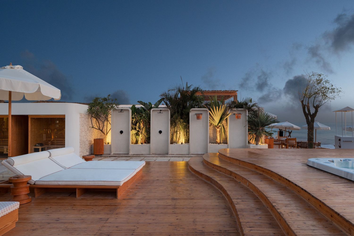 Luxury beachfront terrace at twilight featuring white sun loungers, jacuzzi, tropical plants, and ocean views.