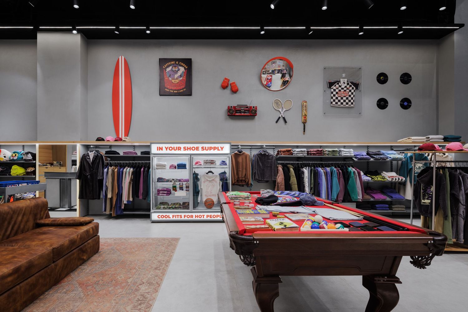 Streetwear boutique interior featuring a red surfboard, pool table merchandise display, clothing racks, and eclectic wall decorations.