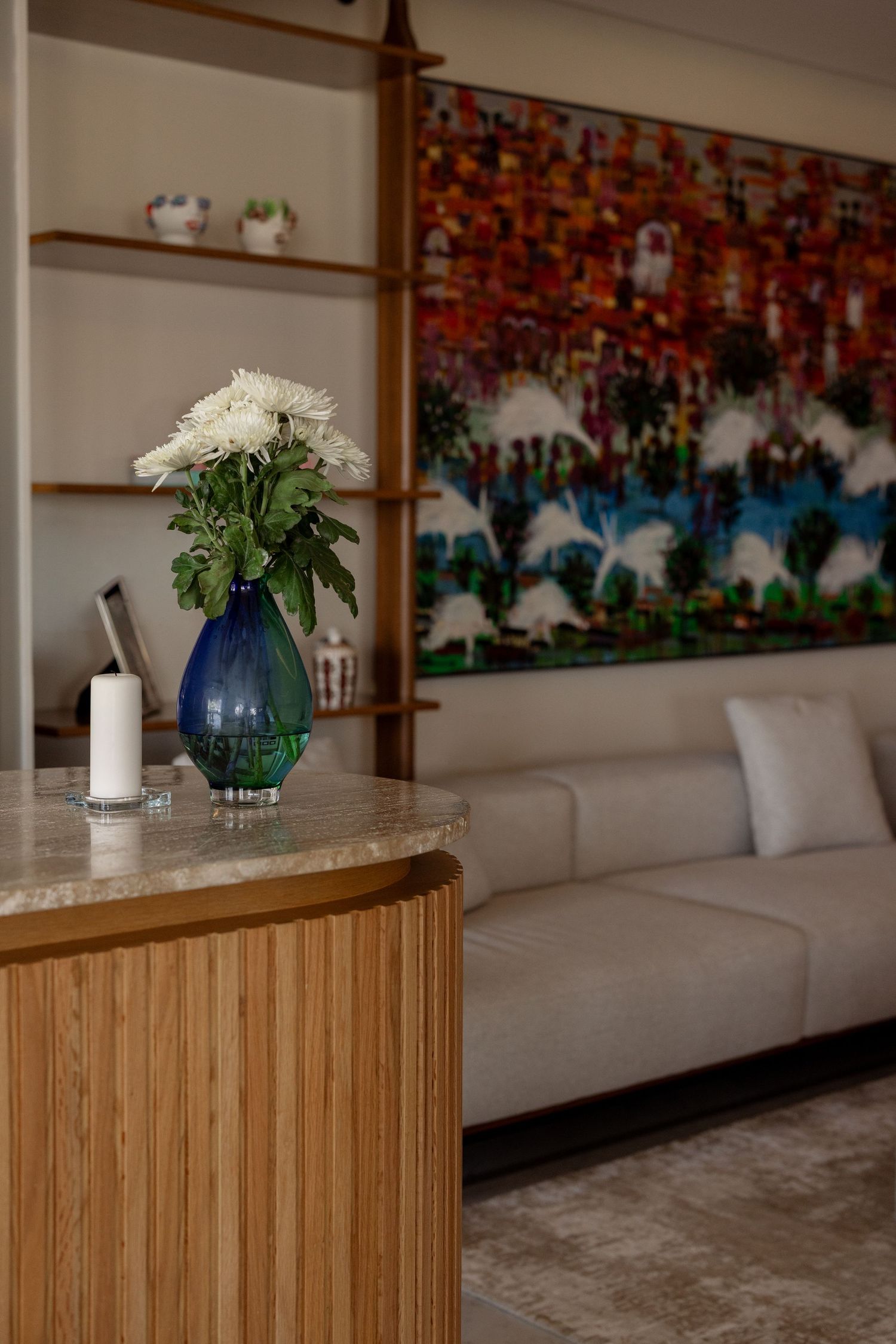 Modern living room with wooden curved bar, blue glass vase with white flowers, and colorful abstract artwork on wall.