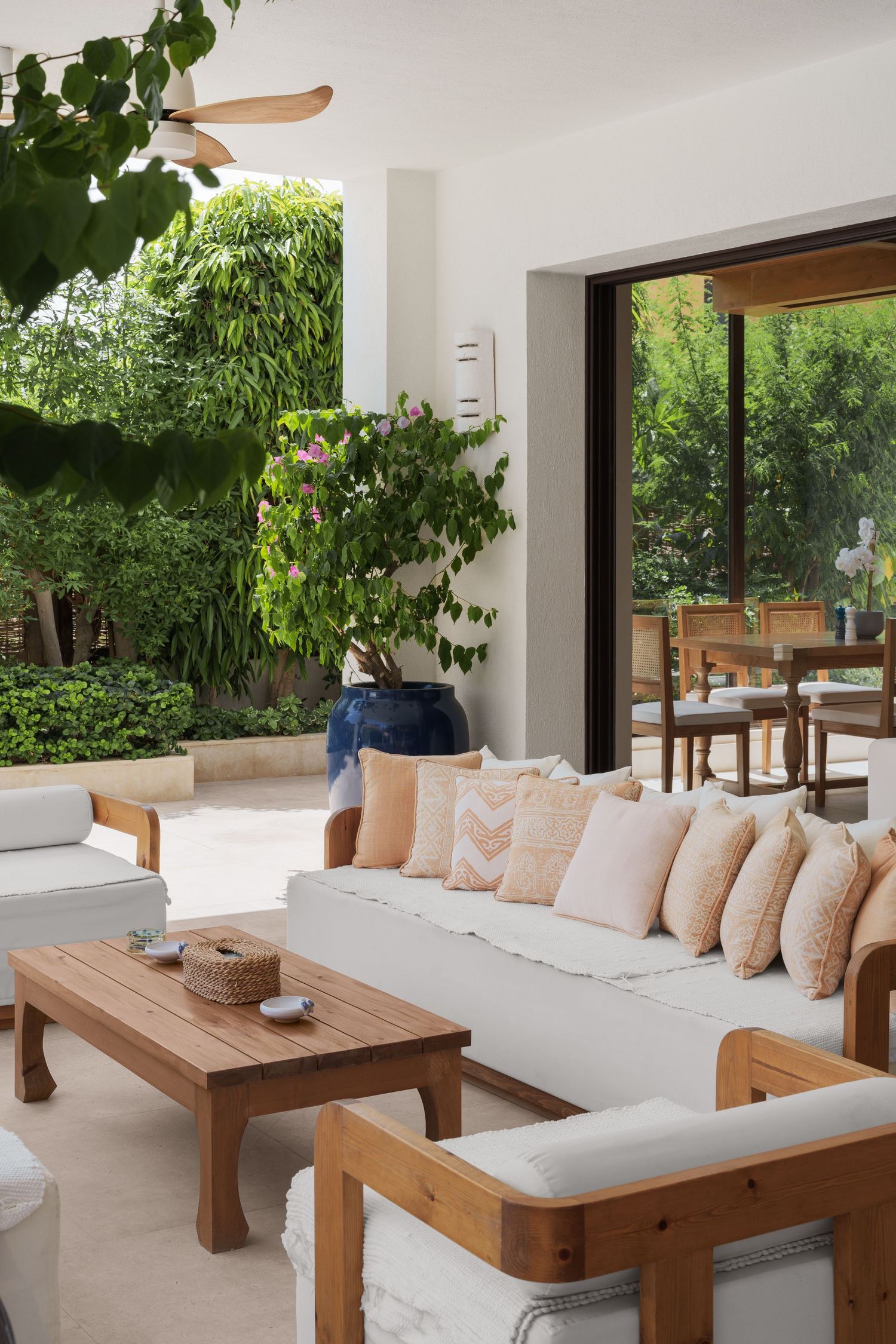 Bright open-air living room with white sofas, wooden furniture, tropical plants, and view to outdoor dining area.