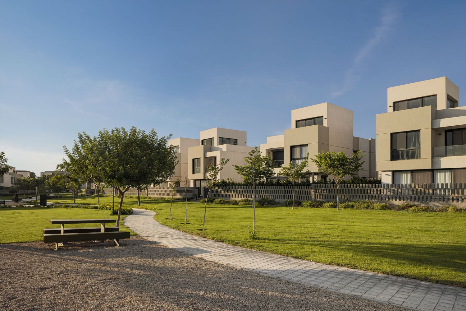 Modern residential complex with cubic beige buildings, lush green lawn, tree-lined pathway, and a wooden bench under clear blue sky.