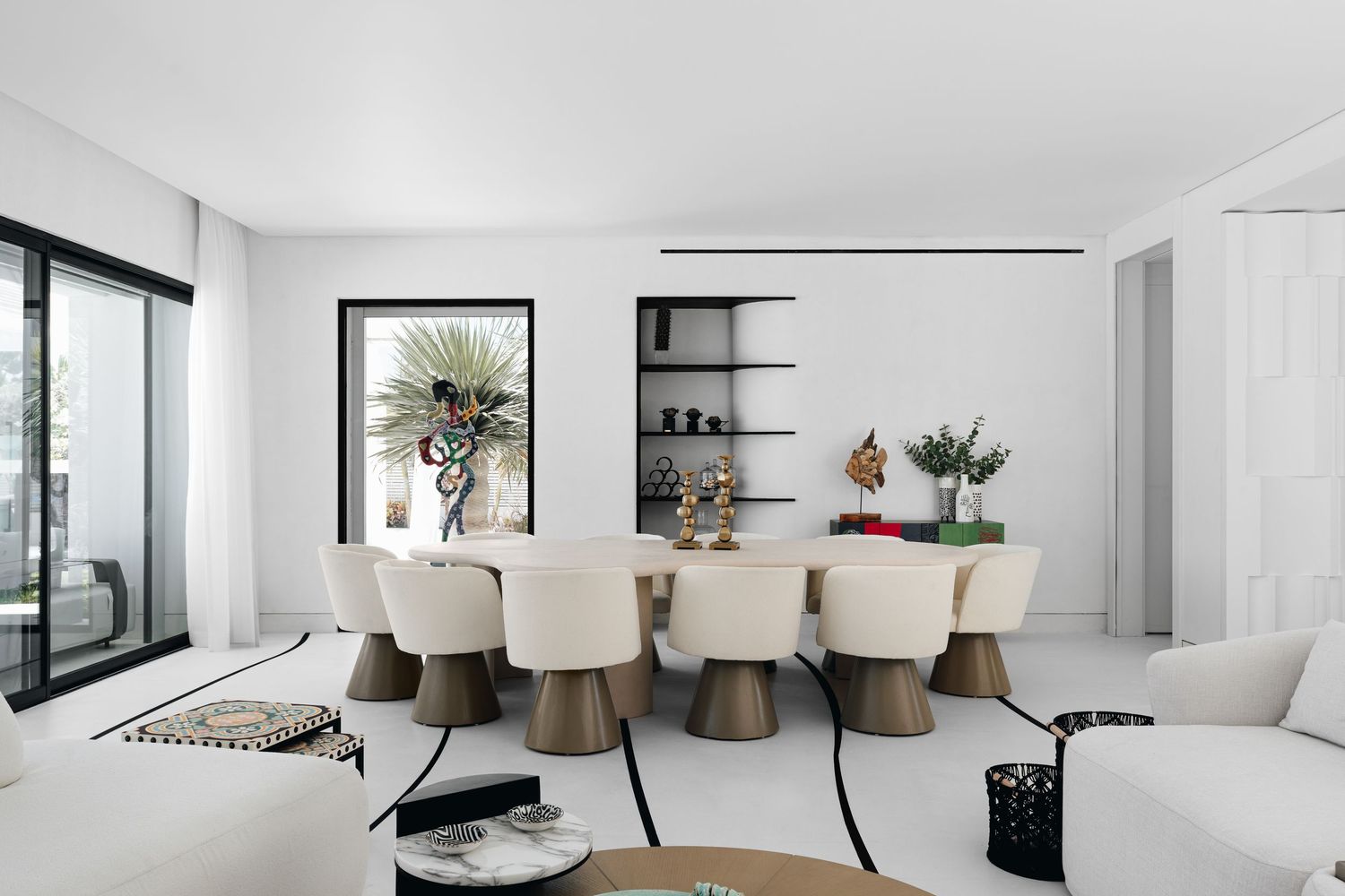 Modern white dining room with cream upholstered chairs, wooden table, built-in shelves, and tropical artwork in a bright space.