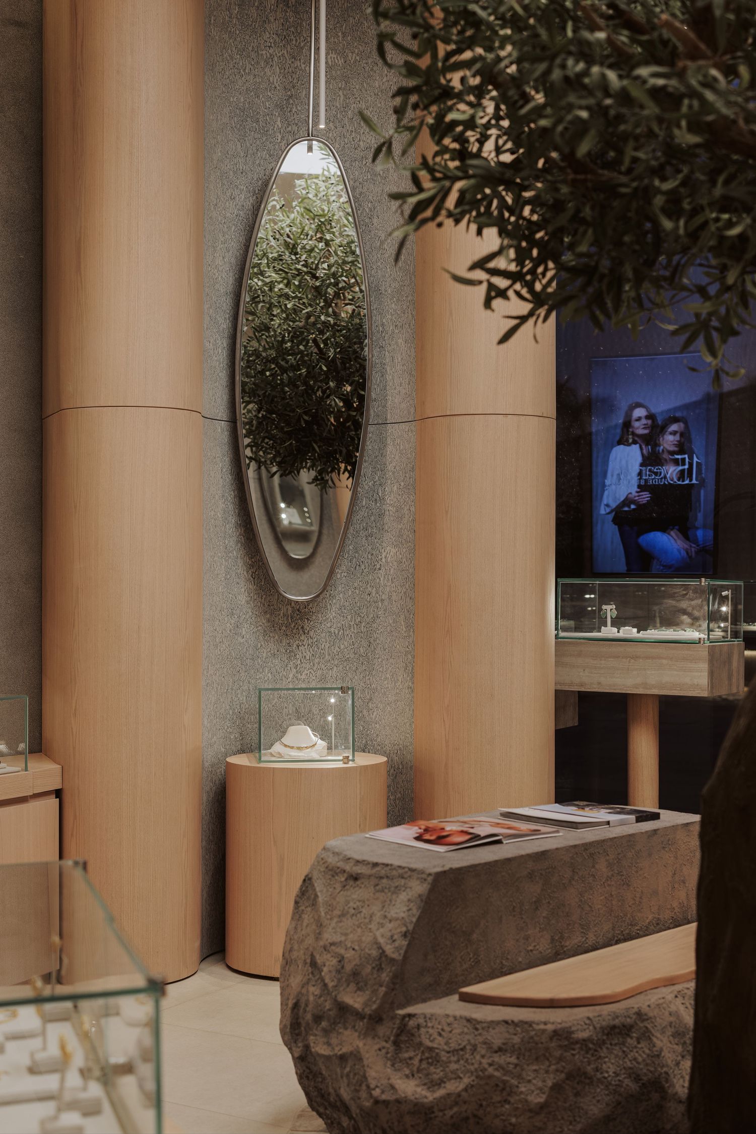 Jewelry boutique interior featuring oval mirror, wood cylindrical columns, stone display table, and olive tree branches.