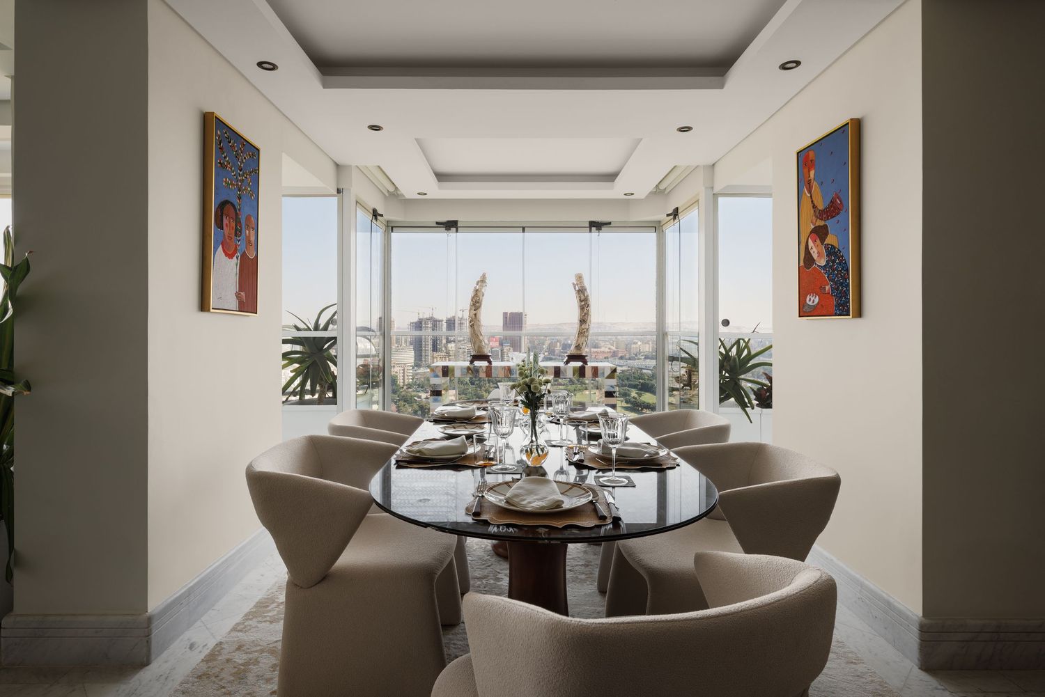 Formal dining room with oval glass table, taupe upholstered chairs, panoramic city views, and vibrant figurative wall art.