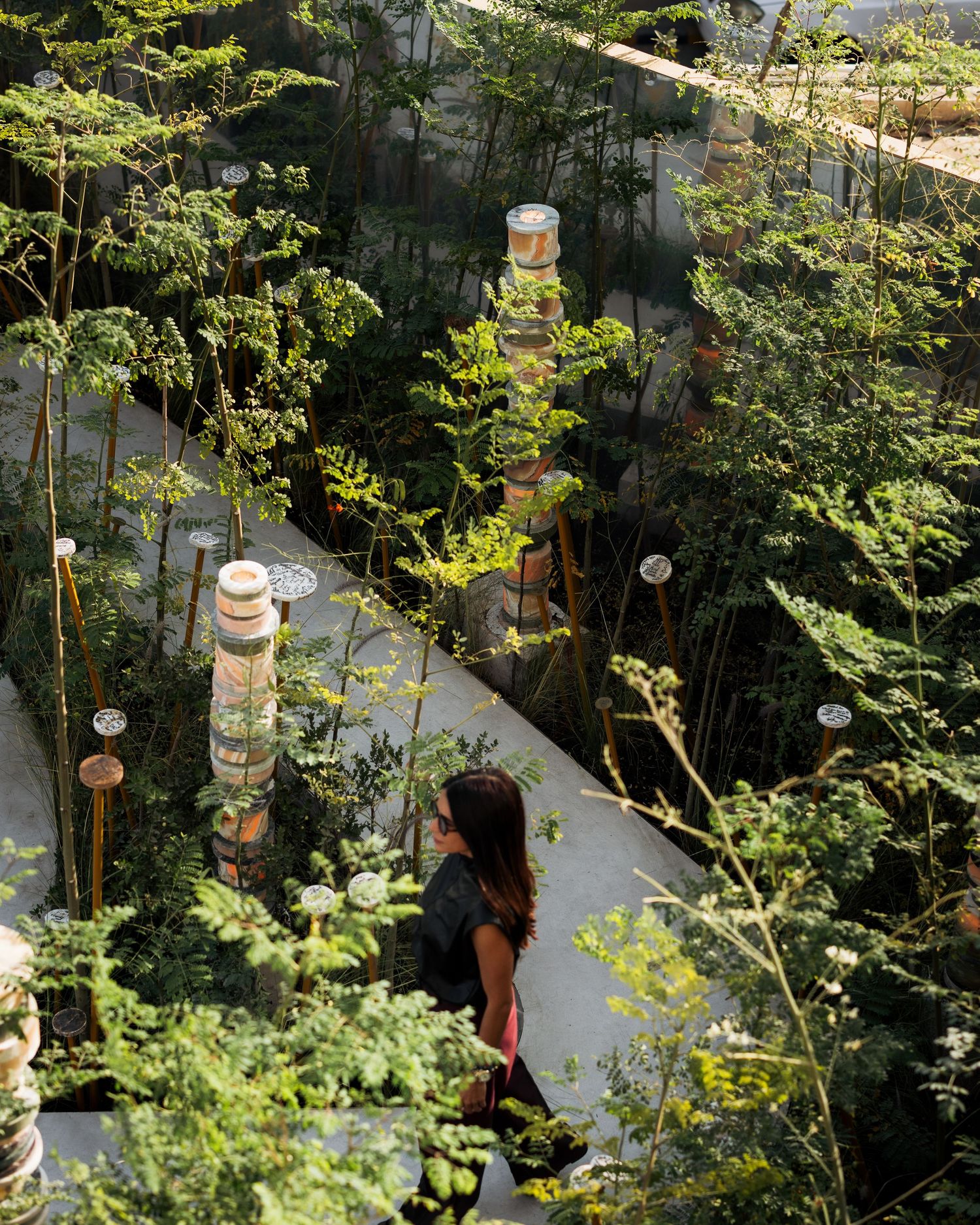 Aerial view of a lush garden installation with a woman walking among tall plants and decorative totem sculptures.